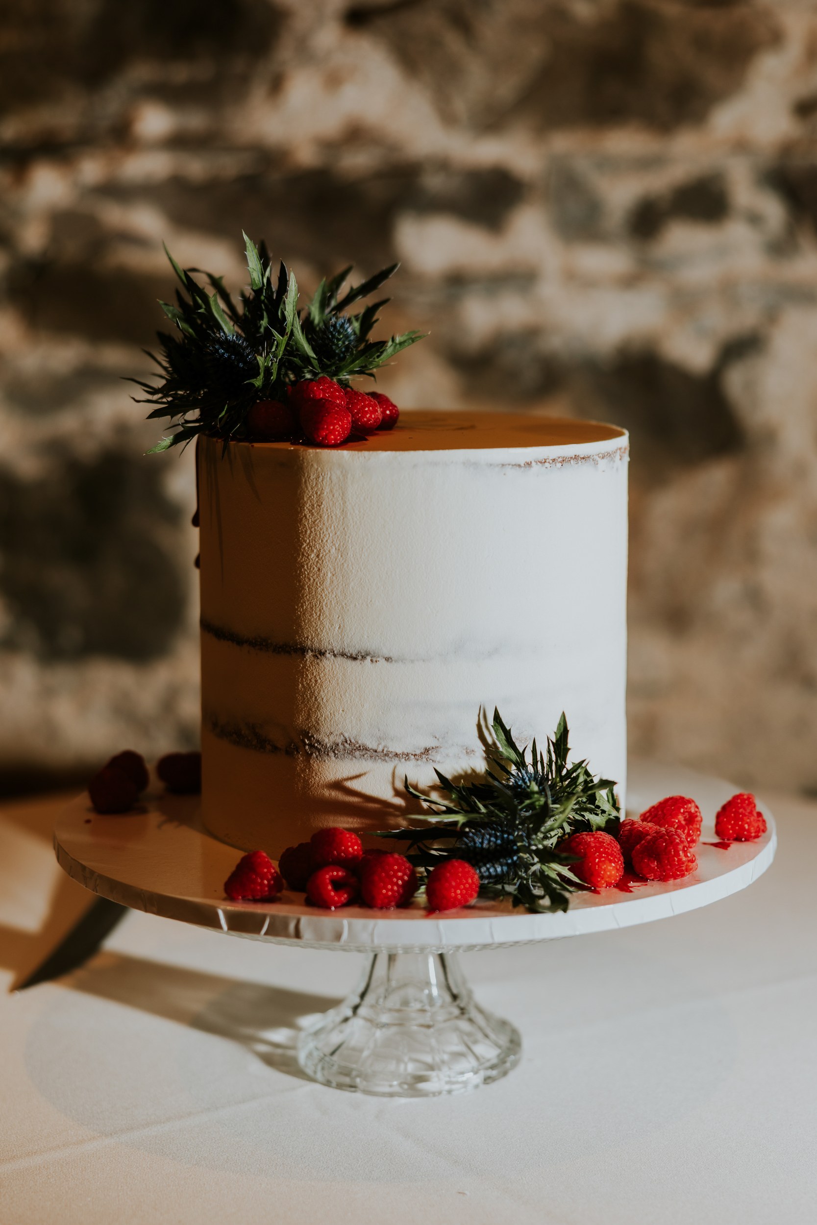 Two-tier white wedding cake decorated with raspberries and blue thistles on a glass stand against a stone wall.
