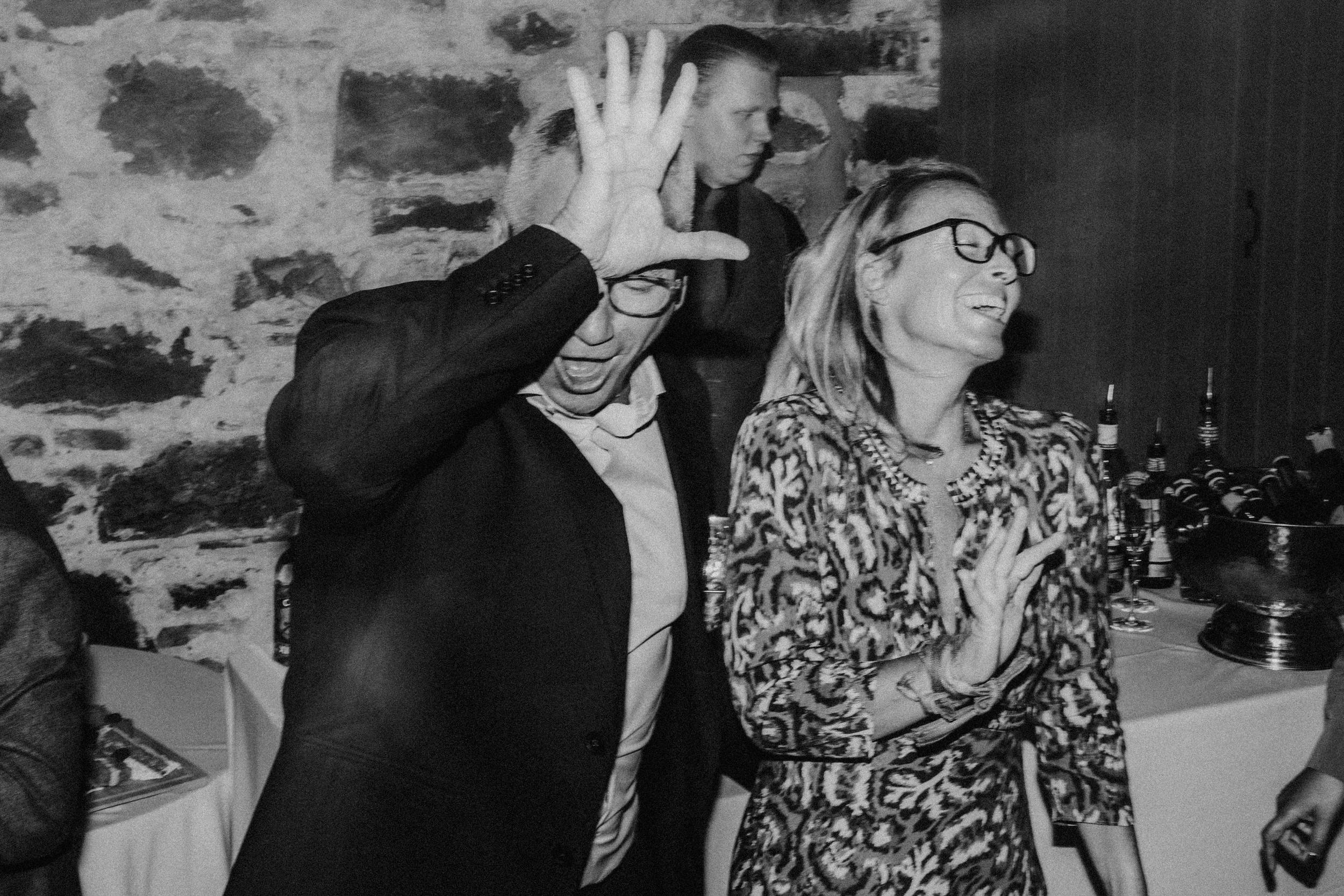 Two wedding guests laughing and dancing by a stone wall, with bottles and wine glasses on a table behind them.
