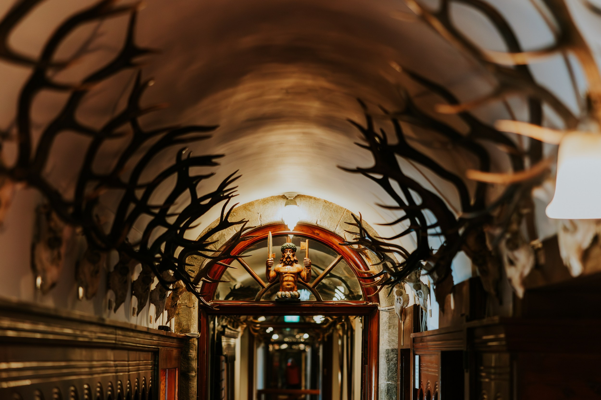 Vaulted corridor lined with mounted antlers and a small carved statue above a wooden door.