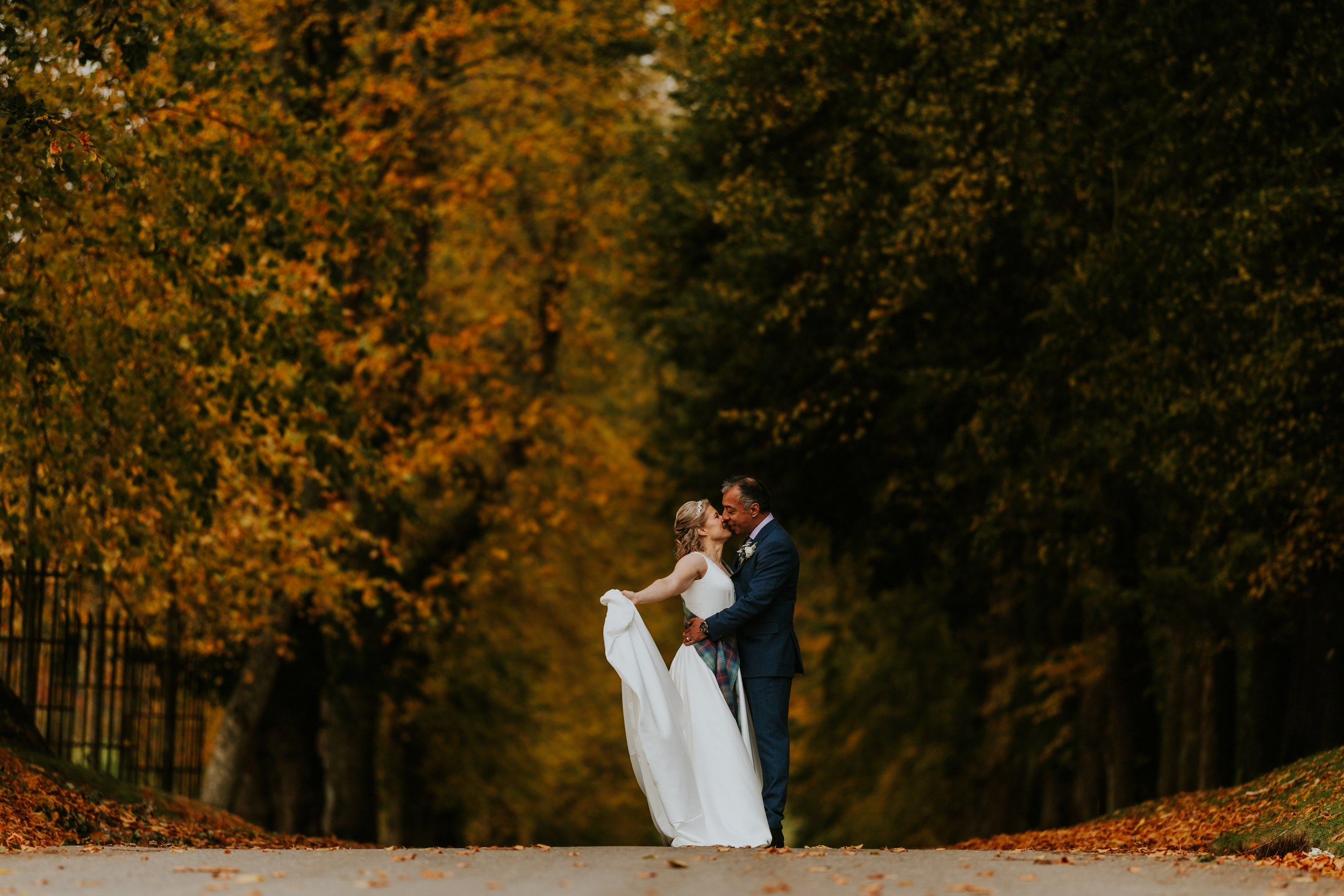 Wedding couple kissing on a tree-lined path with autumn leaves, bride holding her dress.