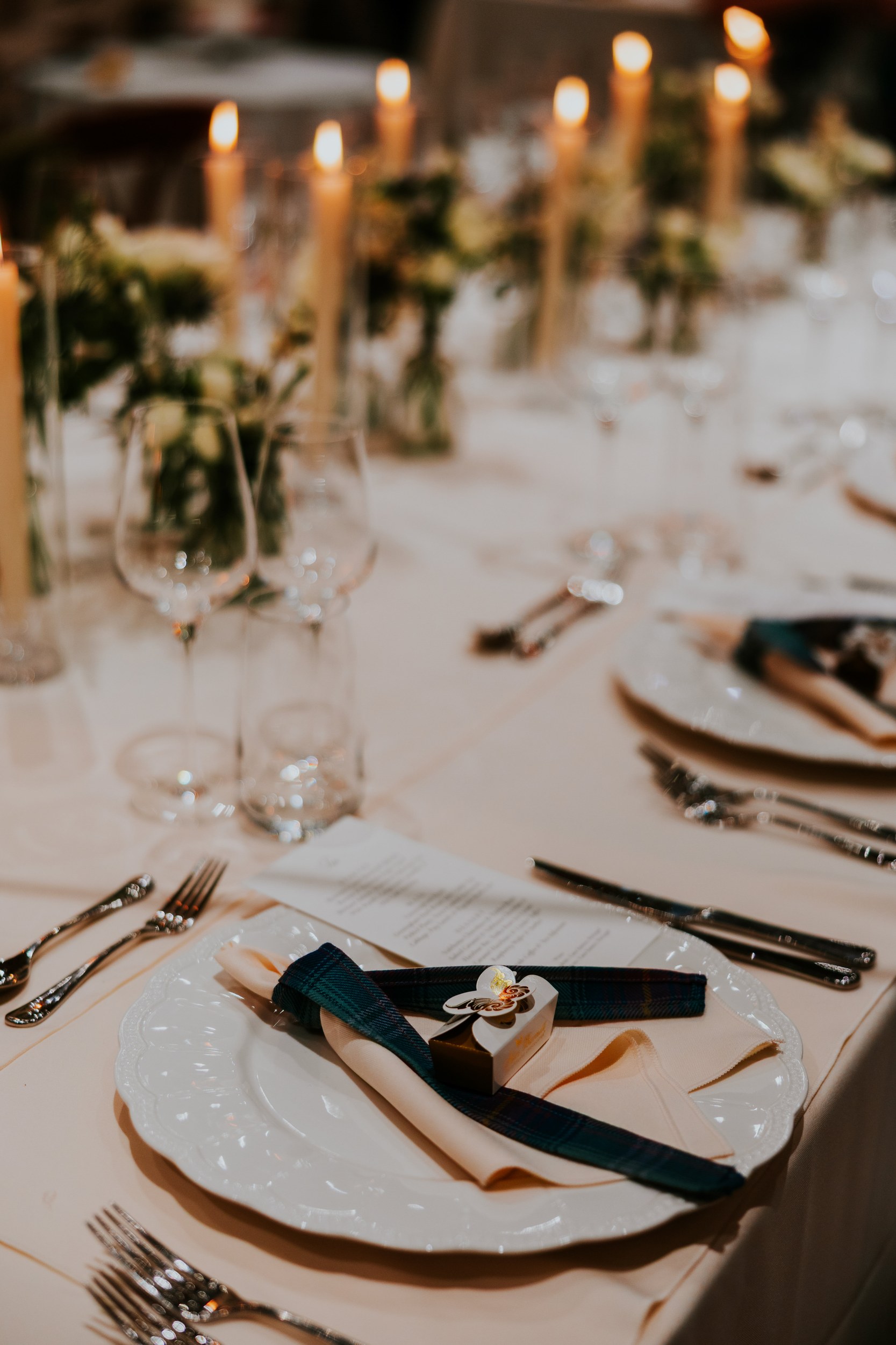Wedding table place setting with white charger, folded napkin tied in tartan and a small favour box, blurred candlelit runner and glasses