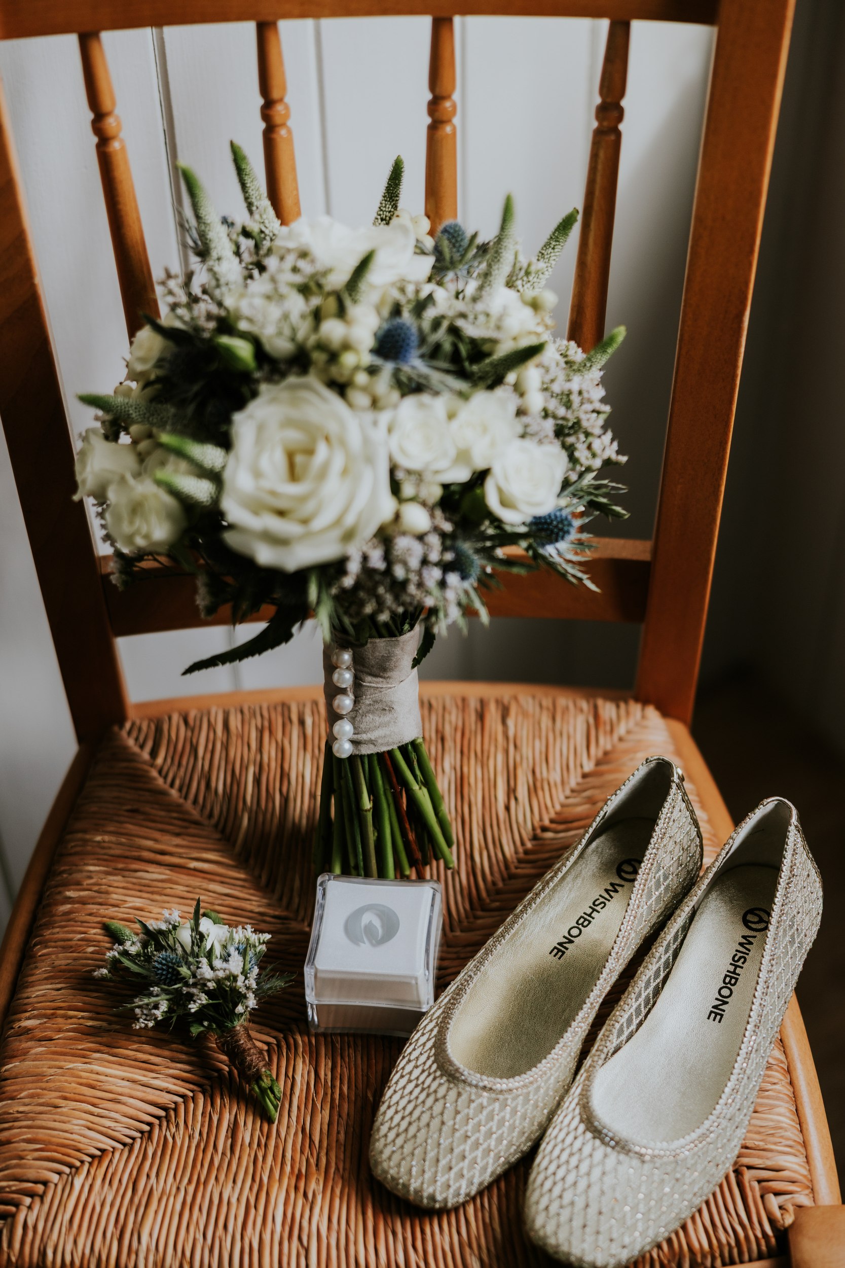 White rose wedding bouquet with thistles on a wooden chair, boutonniere, ring box and pale metallic shoes