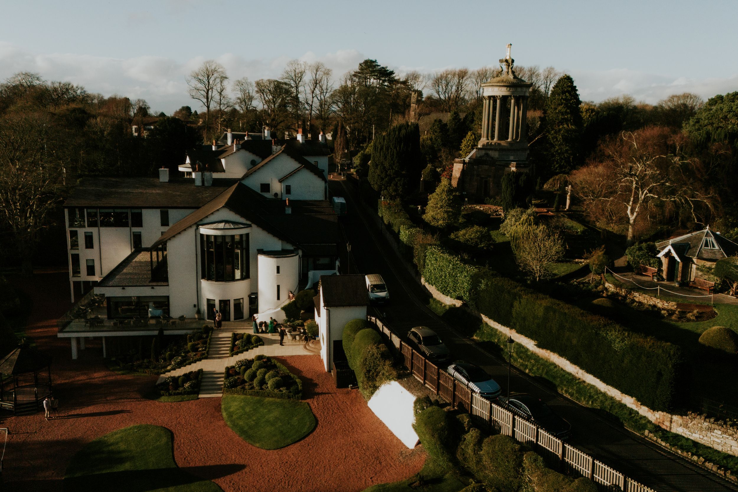 Aerial view of Brig o' Doon House Hotel, guests at the entrance, formal gardens and a nearby memorial column.