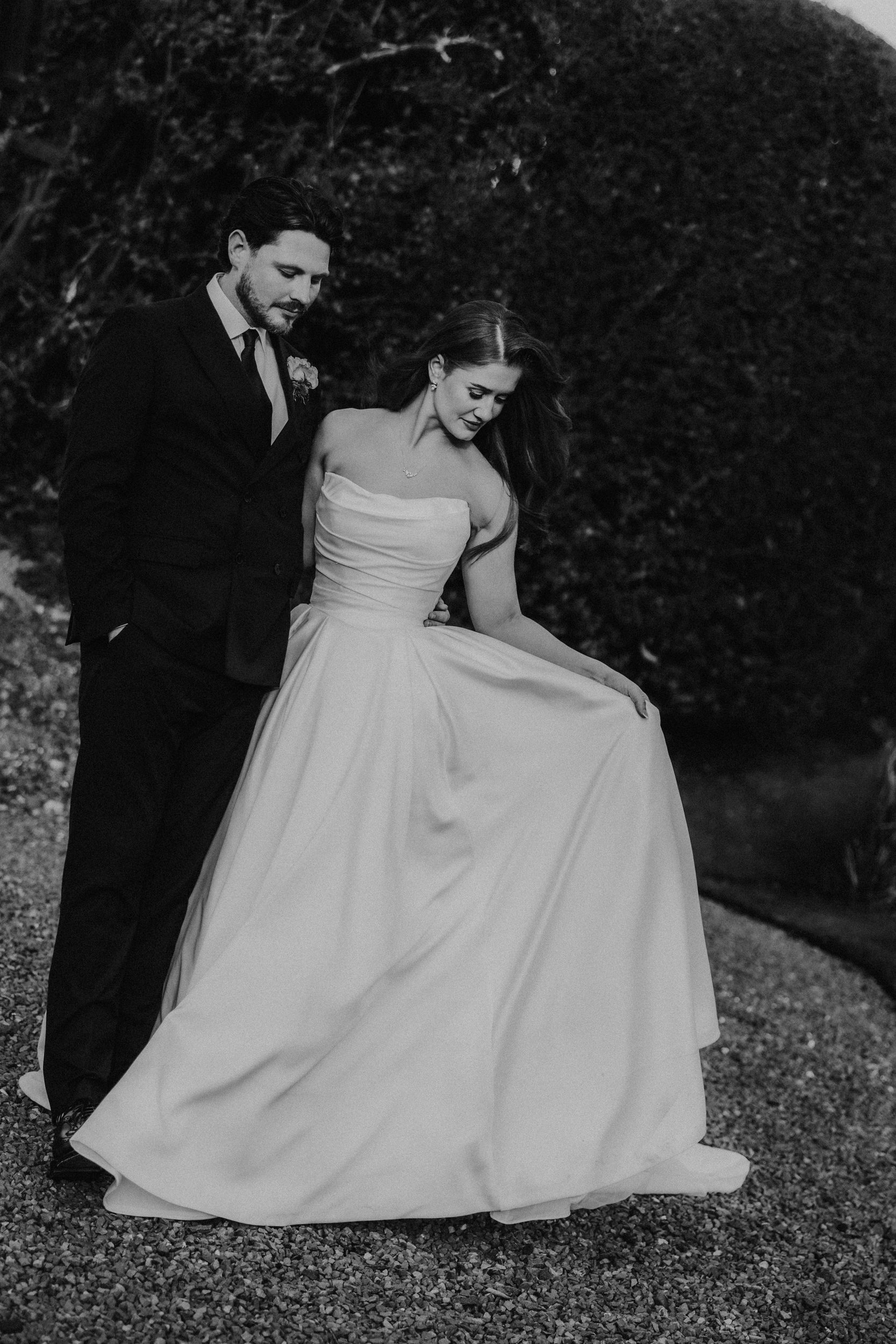 Black-and-white portrait of groom in dark suit beside bride in strapless wedding gown holding her skirt.