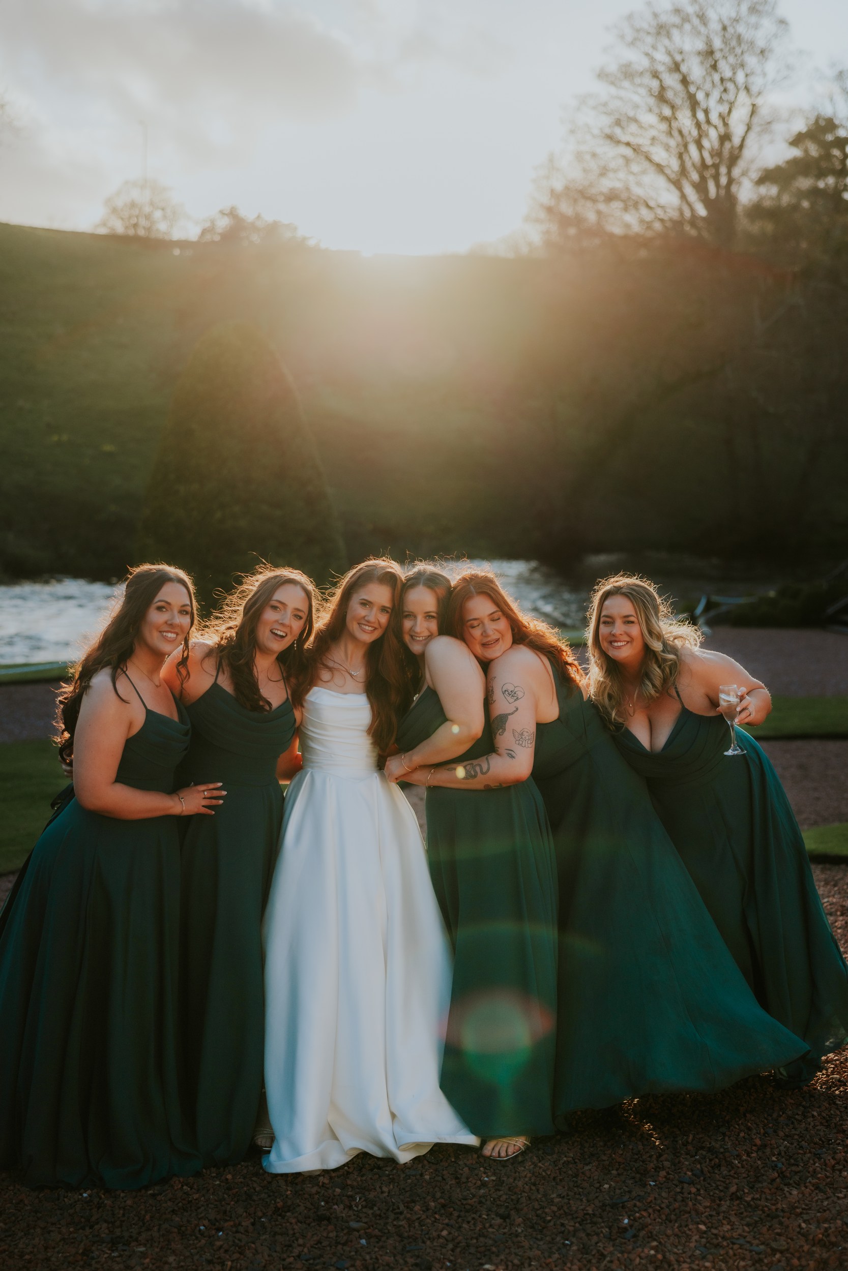 Bride and five bridesmaids in dark green dresses huddled and smiling outdoors at sunset by a river.