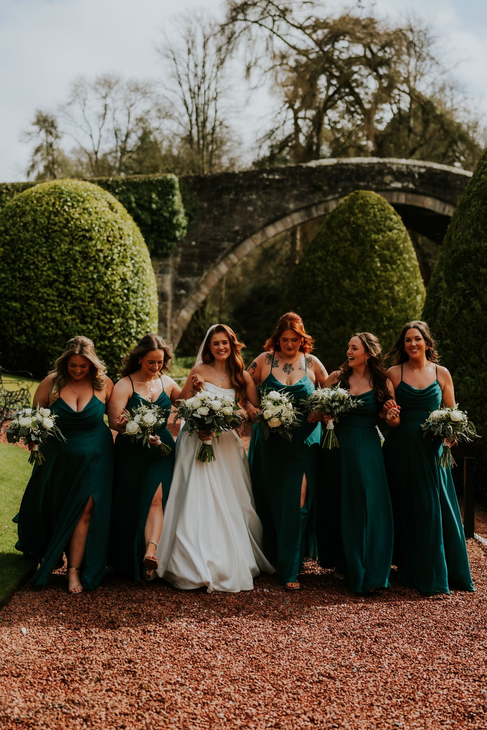 Bride and five bridesmaids in teal dresses holding bouquets, walking past topiary with a stone bridge behind them.