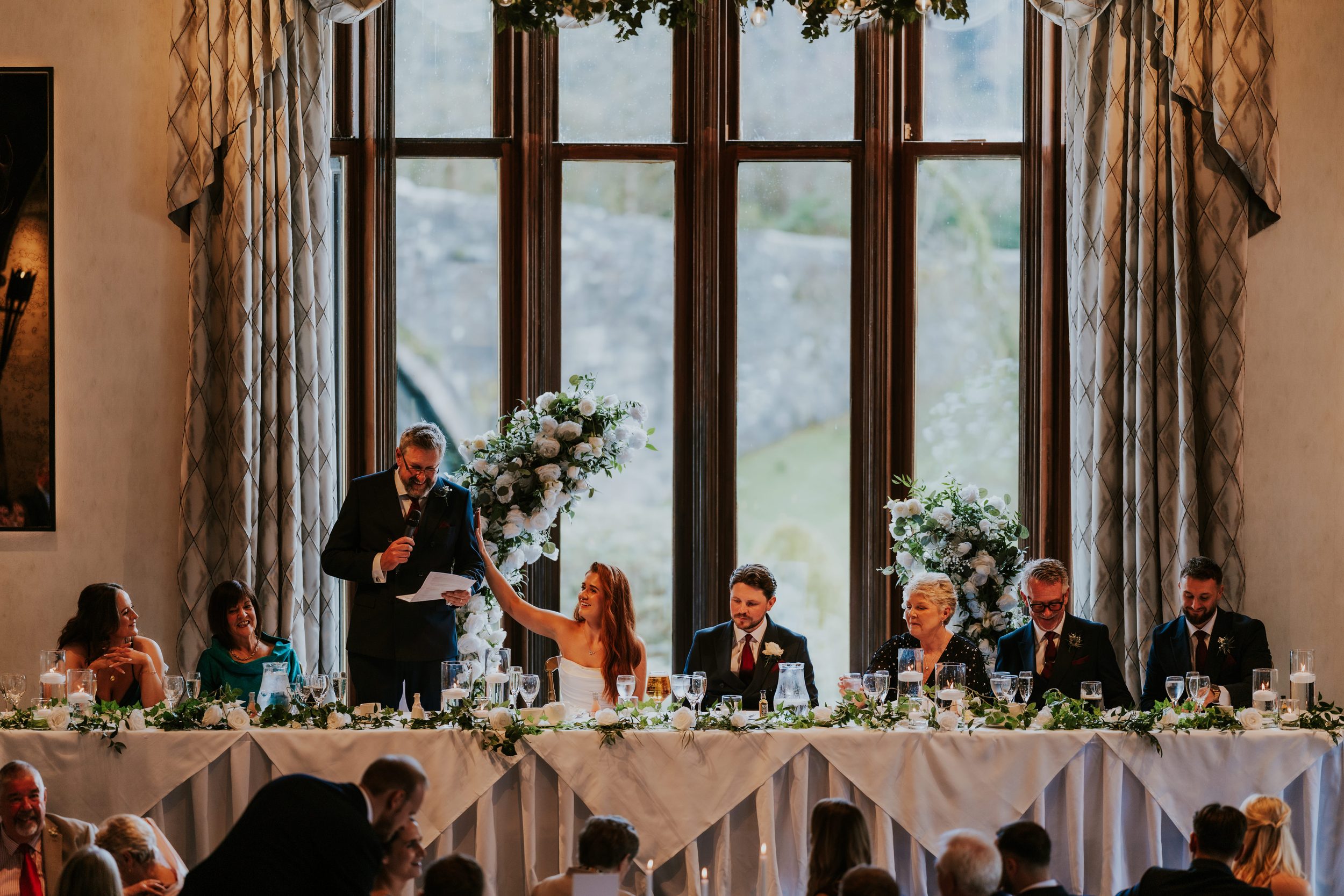 Bride and groom at the head table as a man gives a speech beside large floral displays in front of tall windows.