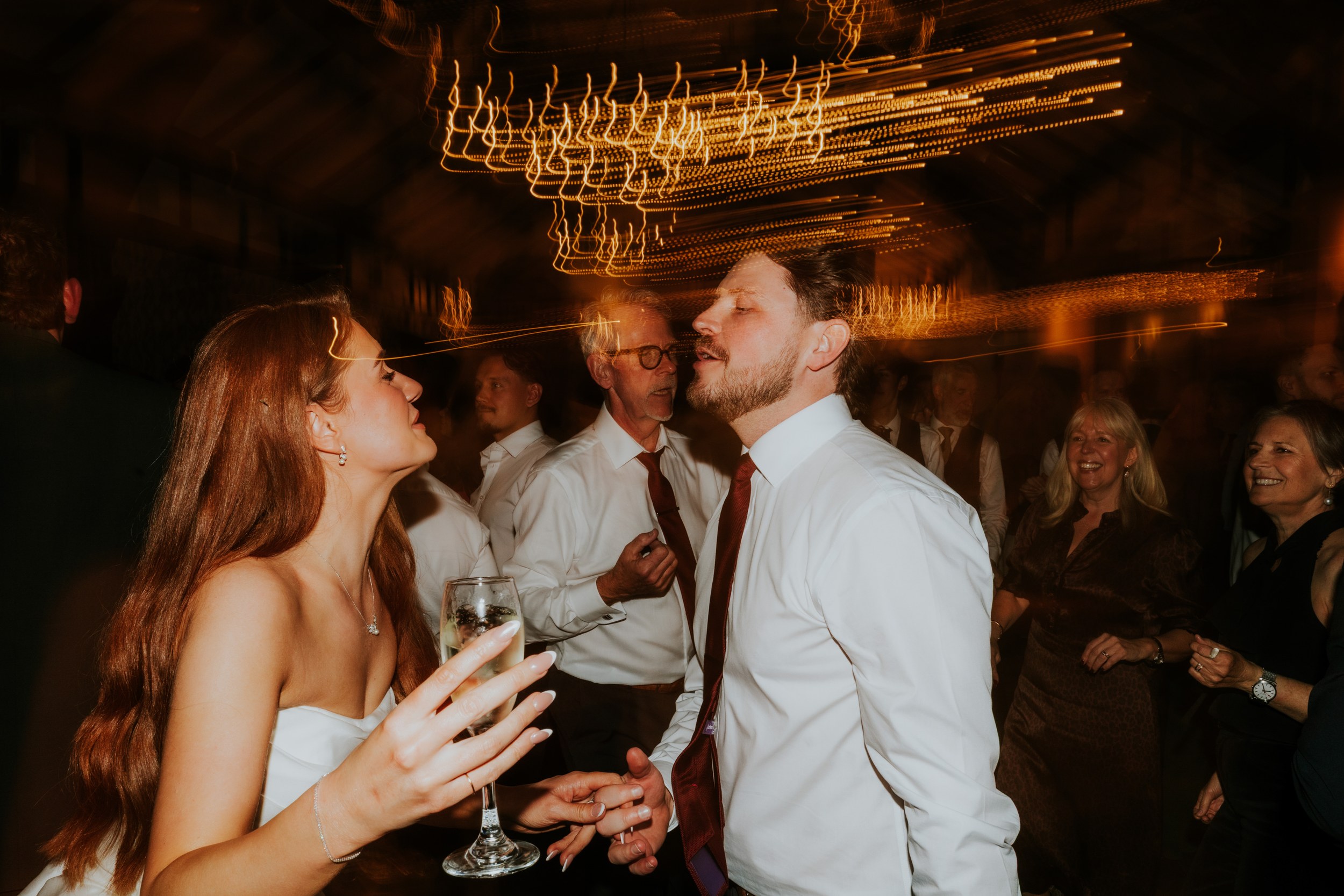 Bride and groom dancing at a reception with warm light trails overhead, guests smiling around them.