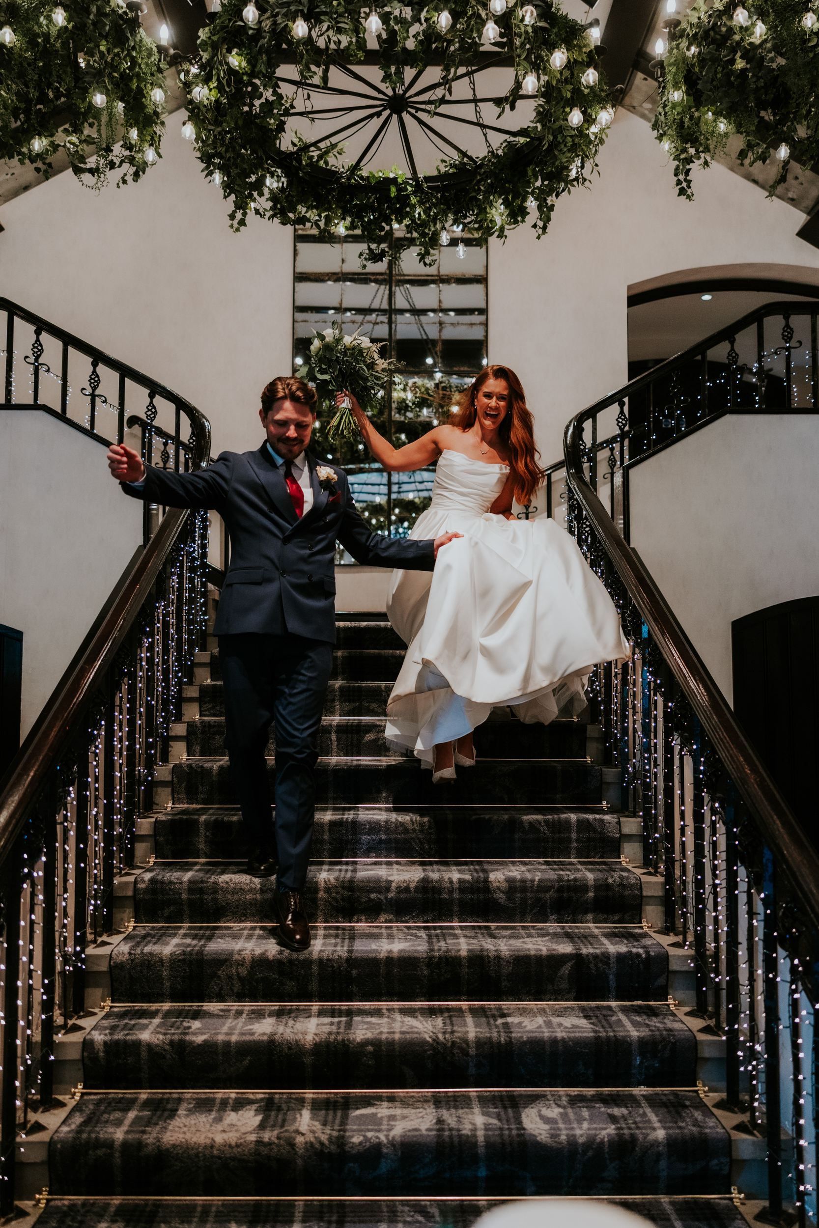 Bride and groom descend a decorated staircase, bride holding bouquet and lifting her dress.