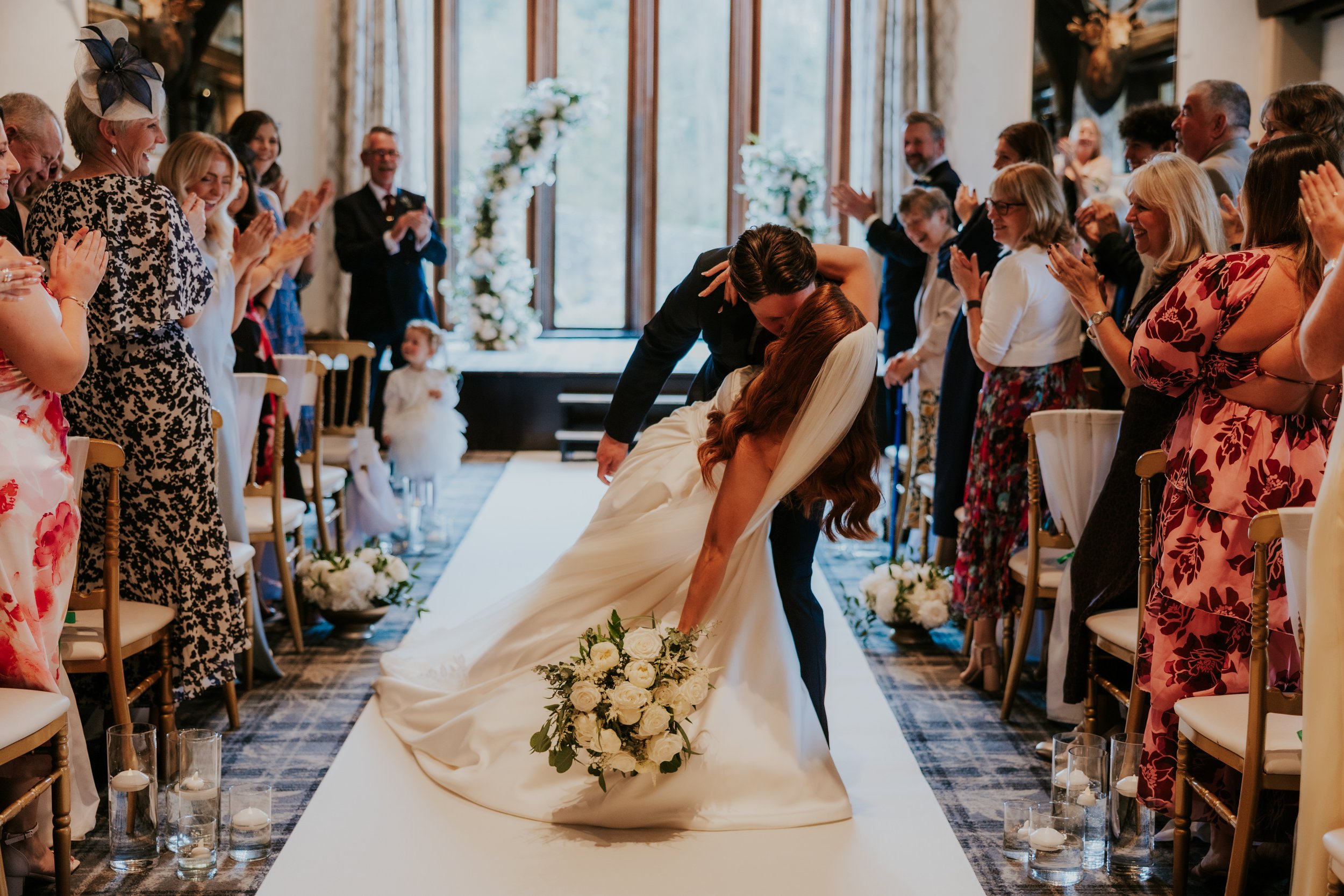 Bride and groom dip for a kiss on the aisle while guests stand and applaud beside floral arrangements.