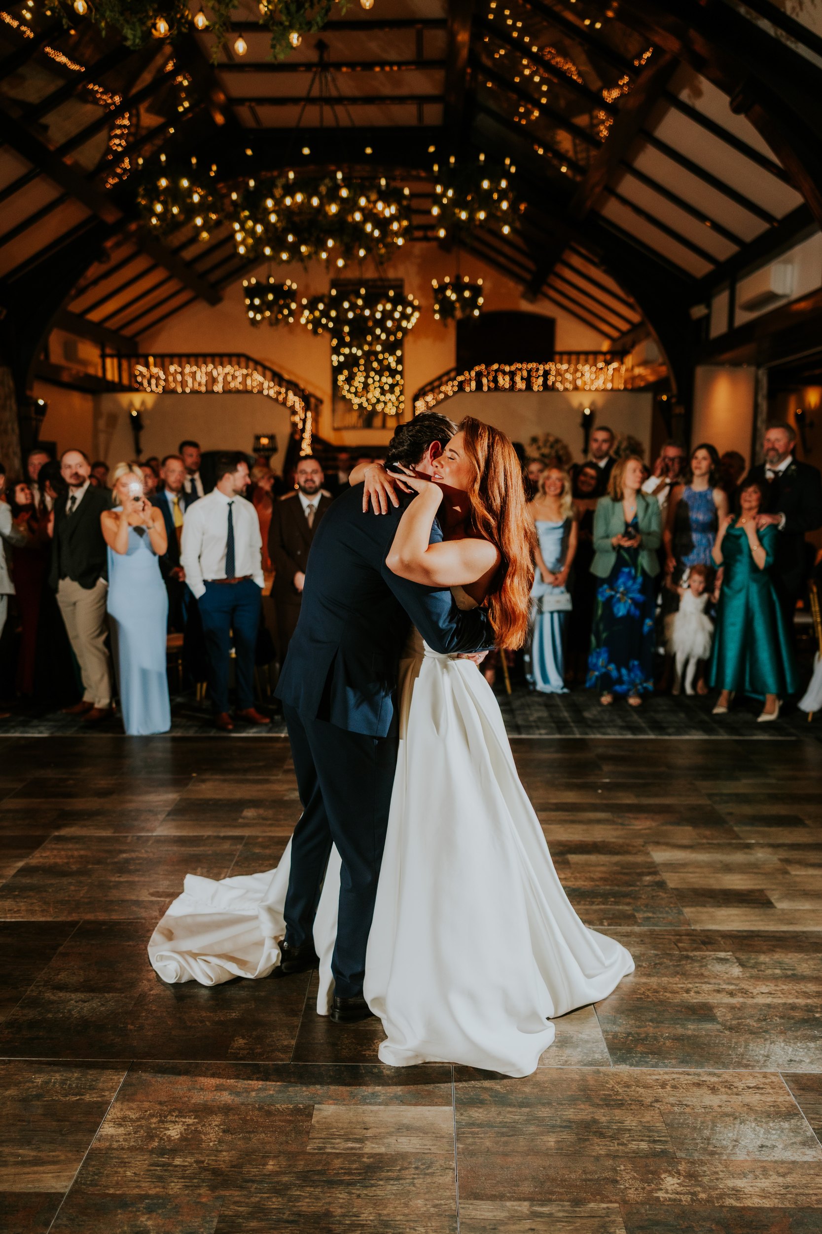 Bride and groom embrace on the dancefloor at their wedding reception, guests watching beneath twinkling lights in a vaulted hall.