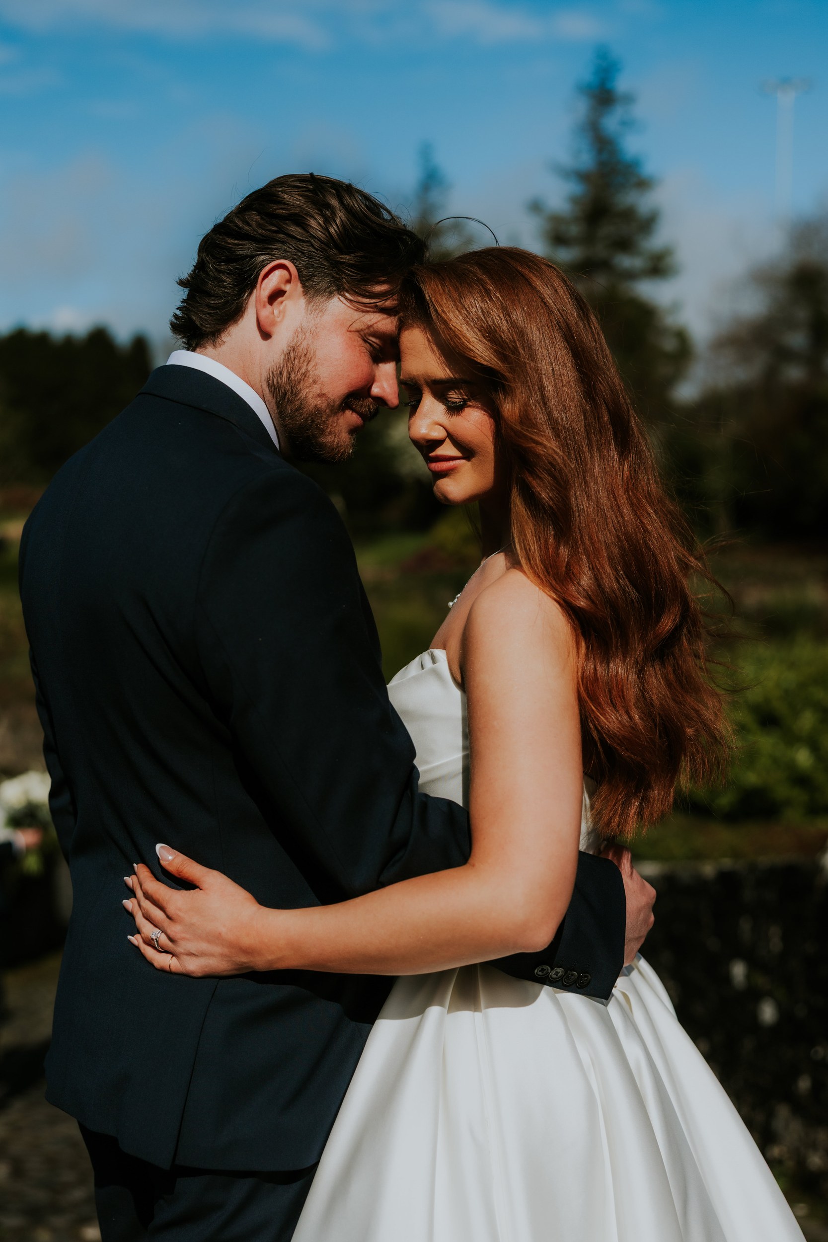 Bride and groom embracing outdoors, foreheads touching; bride in a strapless white gown and groom in a dark suit.