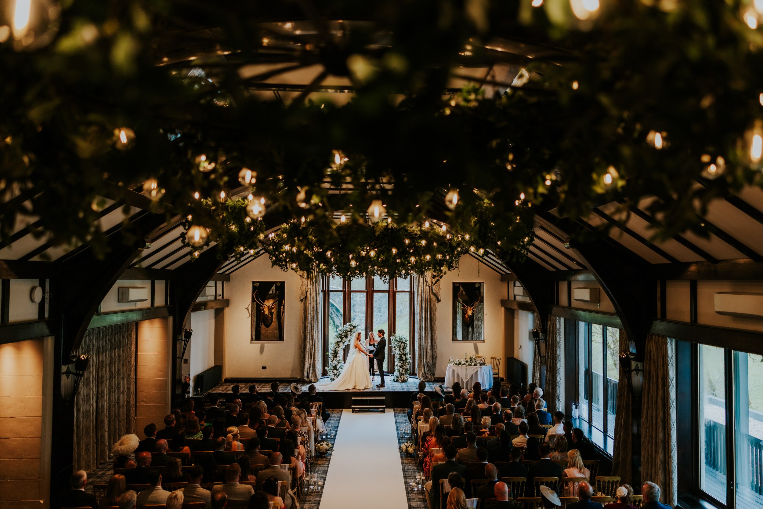 Bride and groom exchanging vows beneath a floral arch in a high‑ceilinged ceremony room with hanging foliage lights.