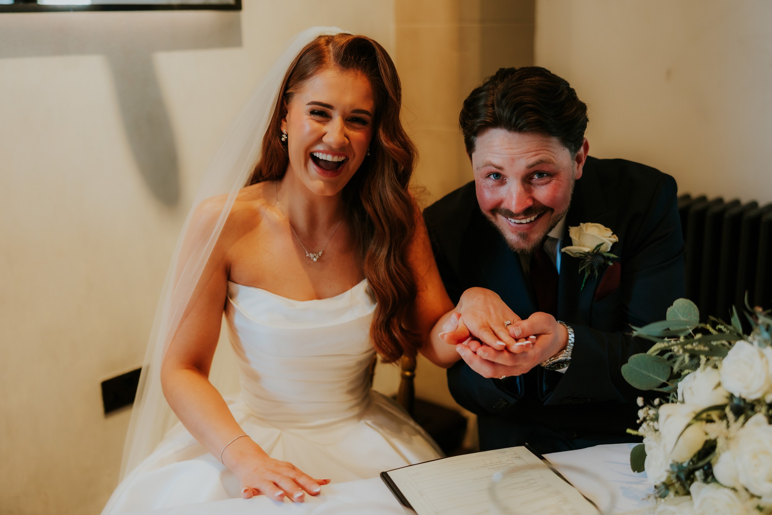 Bride and groom laughing at signing table, bride in veil and strapless gown, groom in dark suit showing her ring.