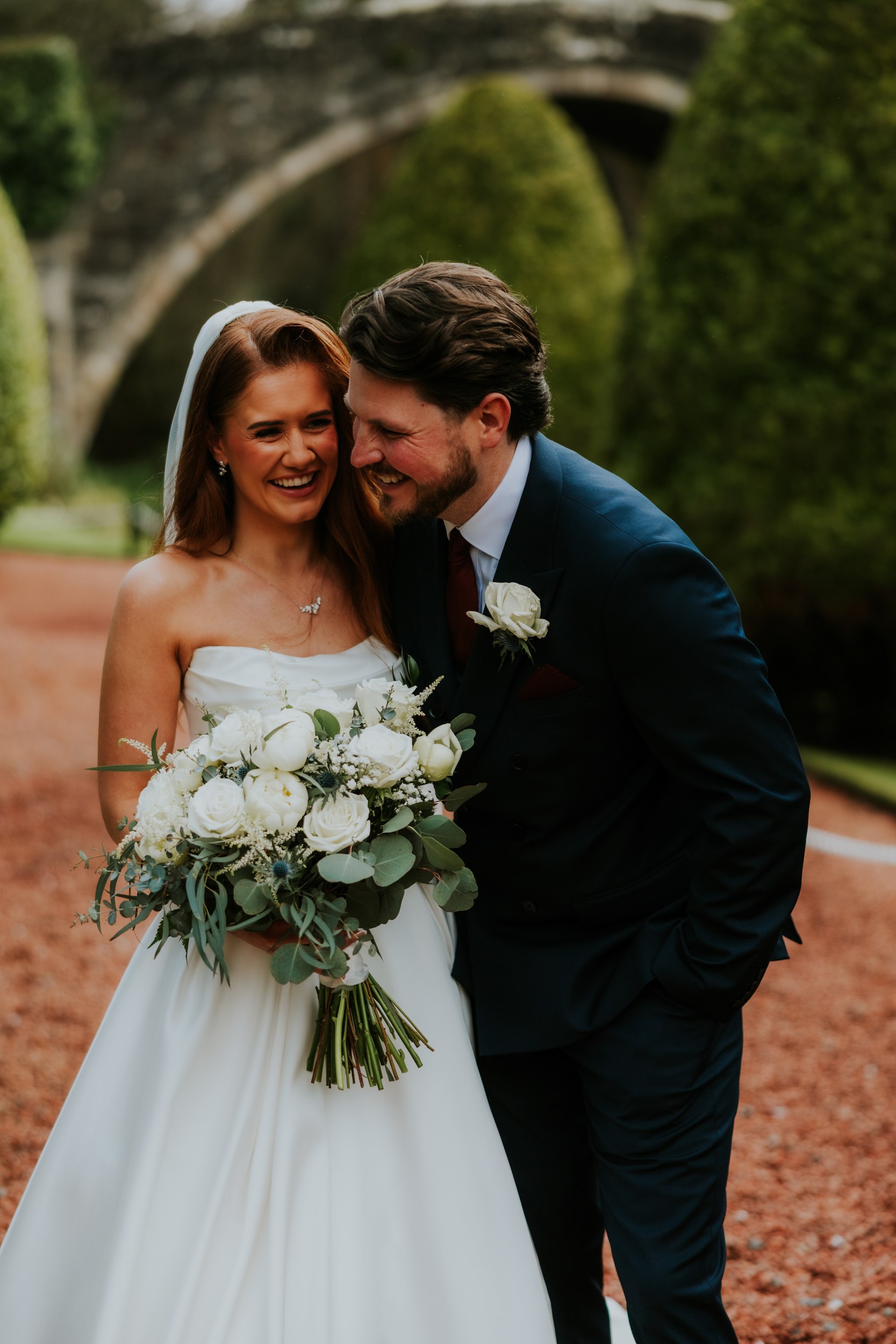 Bride and groom laughing, bride holding white rose bouquet with stone arch and yew trees behind them.