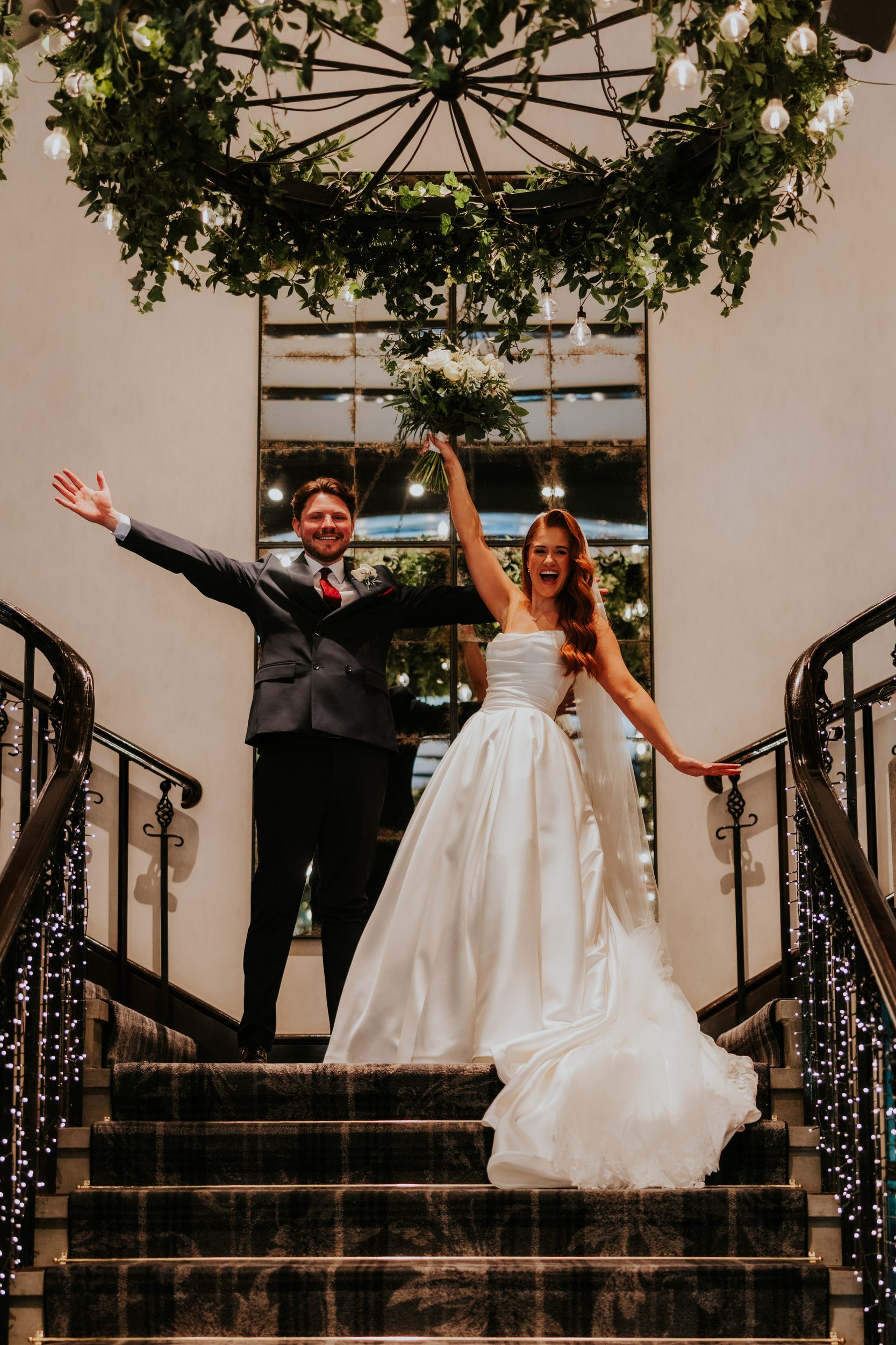 Bride and groom on a staircase, bride raises bouquet overhead and both cheer beneath hanging greenery.