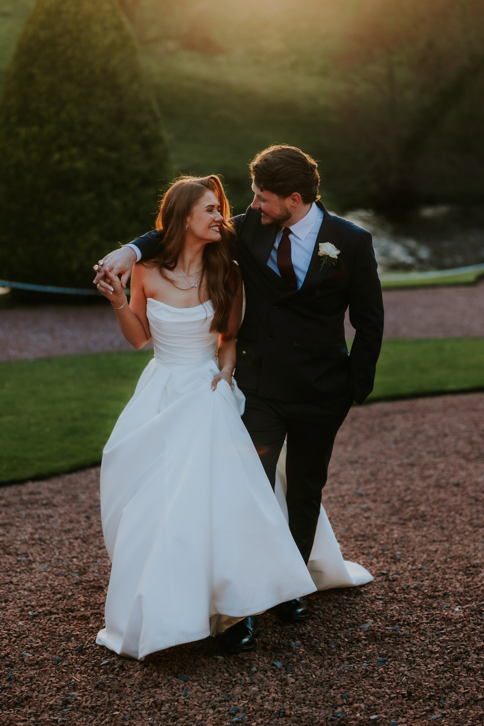 Bride and groom walk arm-in-arm on a gravel path, smiling in golden-hour light.