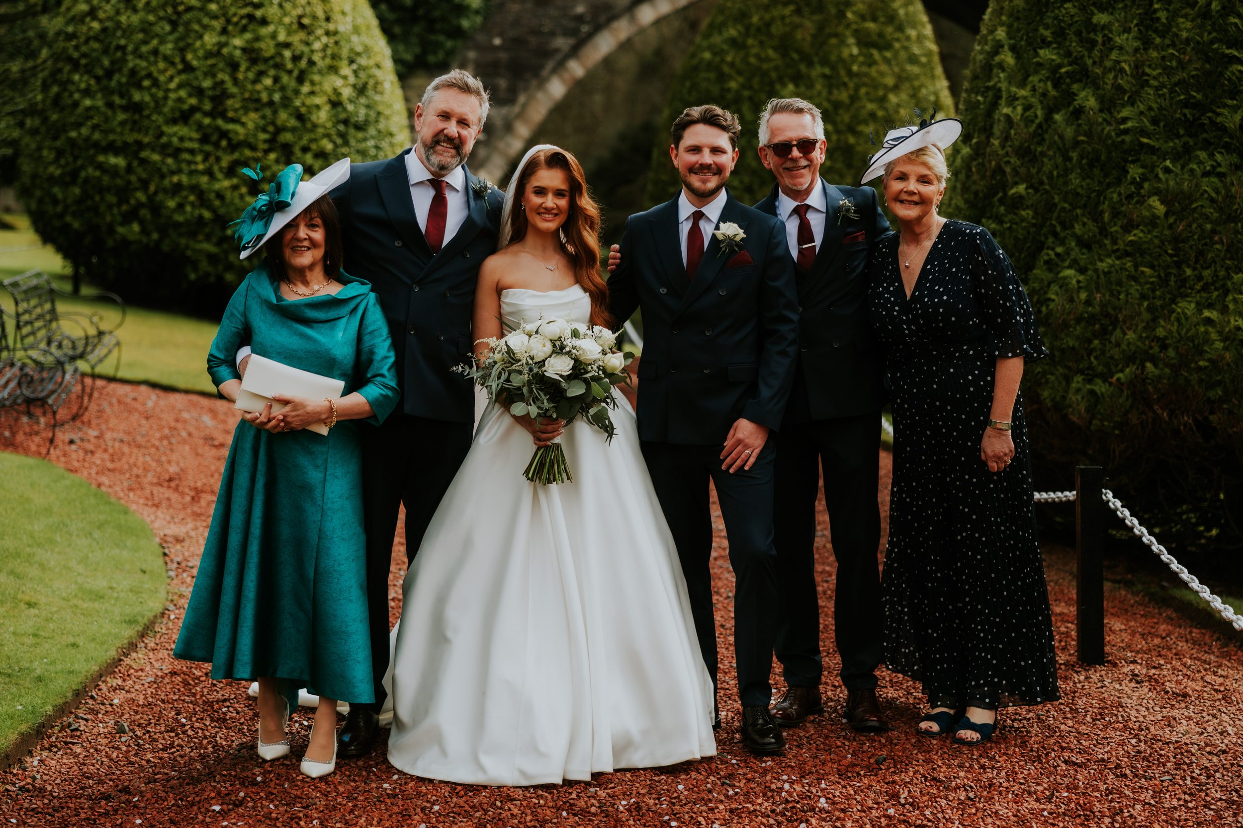Bride and groom with four relatives posed outdoors; bride holds white bouquet, men in dark suits with burgundy ties, two women wearing hats.