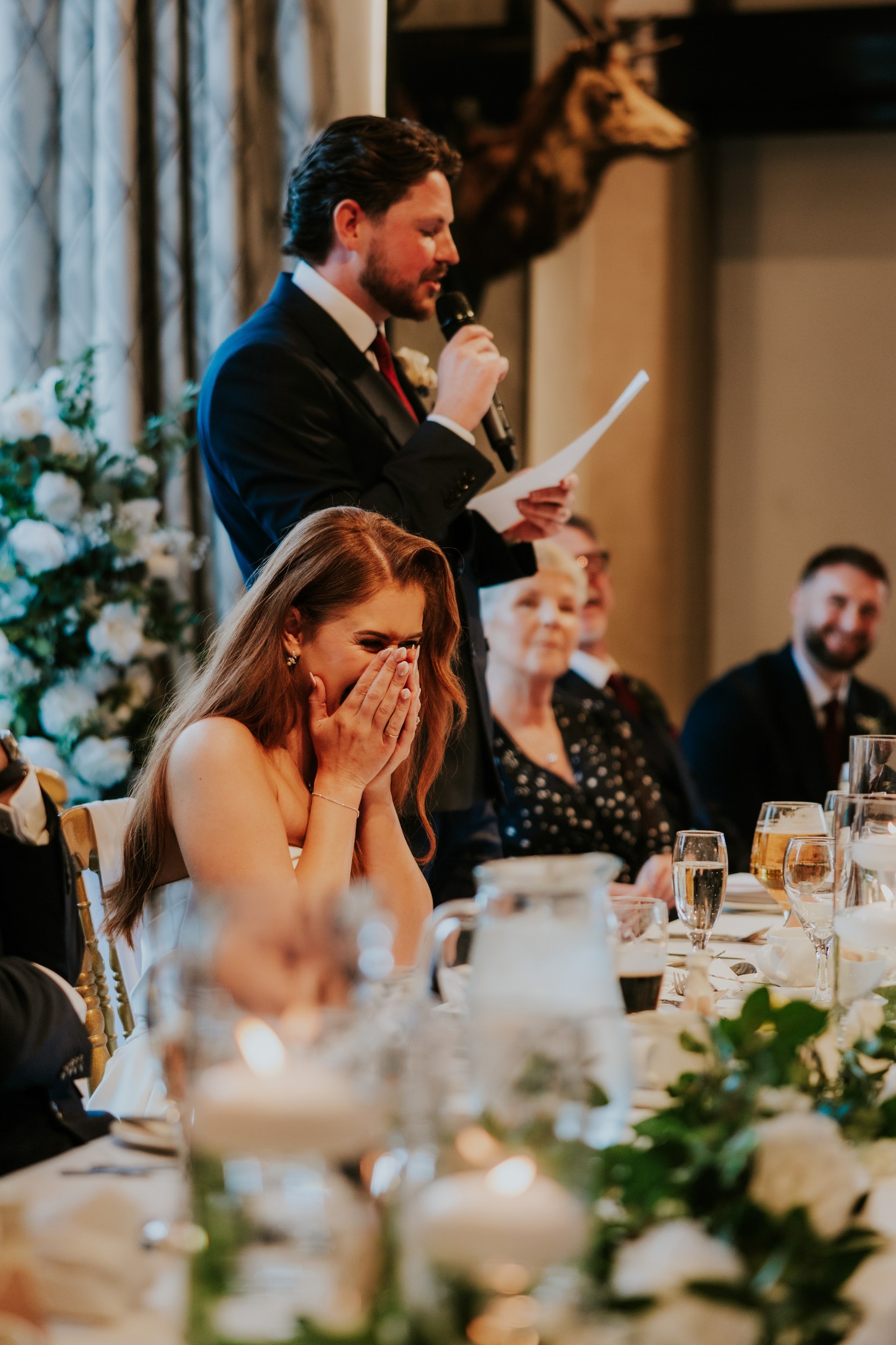 Bride covering her face laughing as a man gives a speech at the wedding reception, table with glasses and flowers in the foreground.