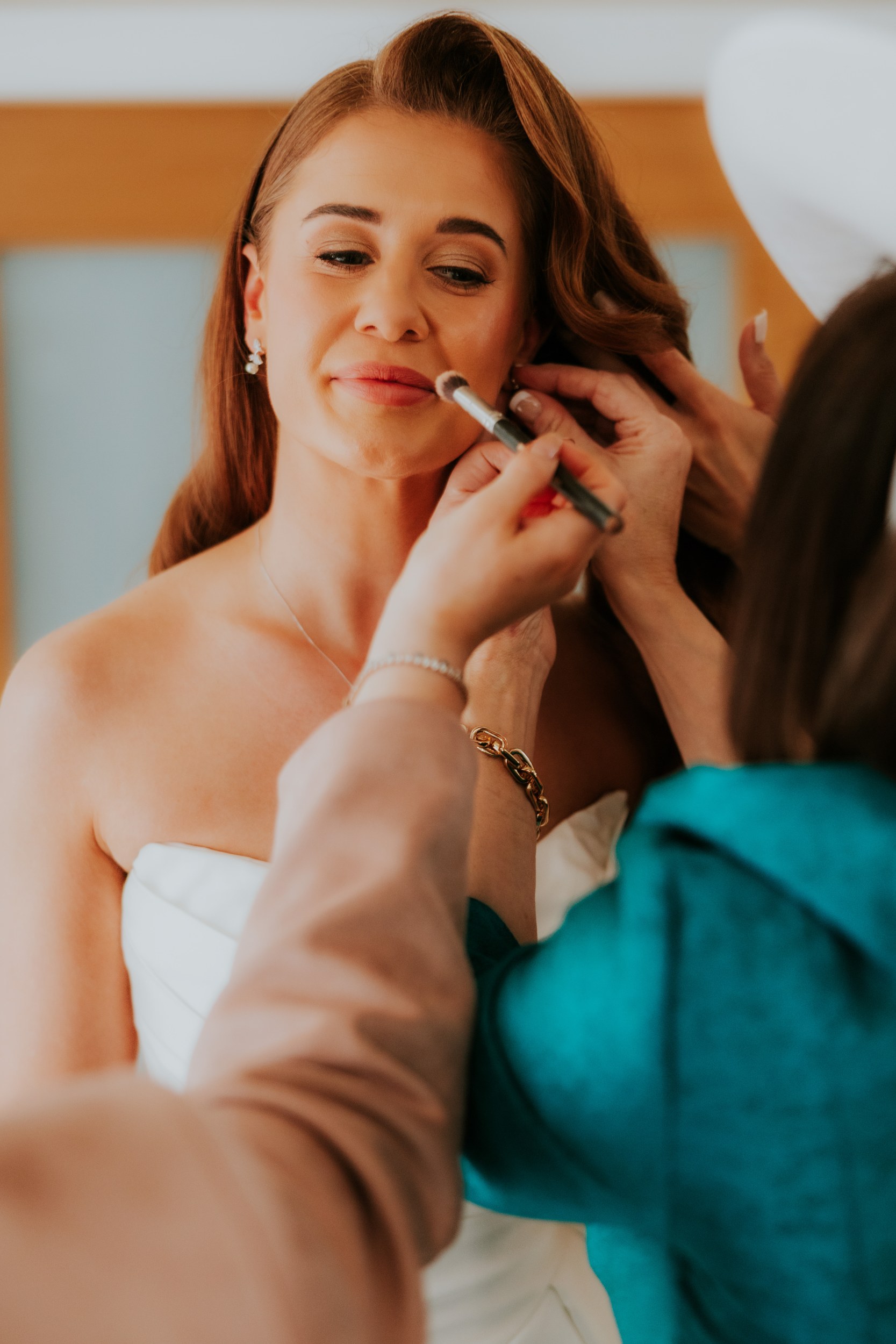 Bride having makeup brushed on and attendants adjusting her earring and hair, close-up of her face and strapless dress.