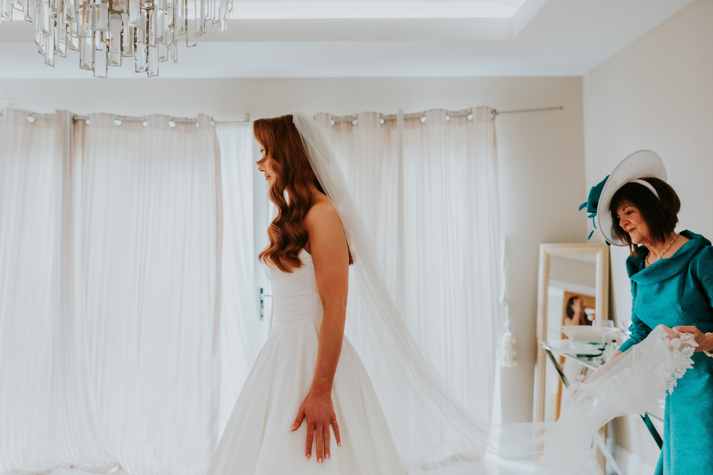 Bride in a strapless dress and long veil in profile as a woman in teal and a hat adjusts her veil.
