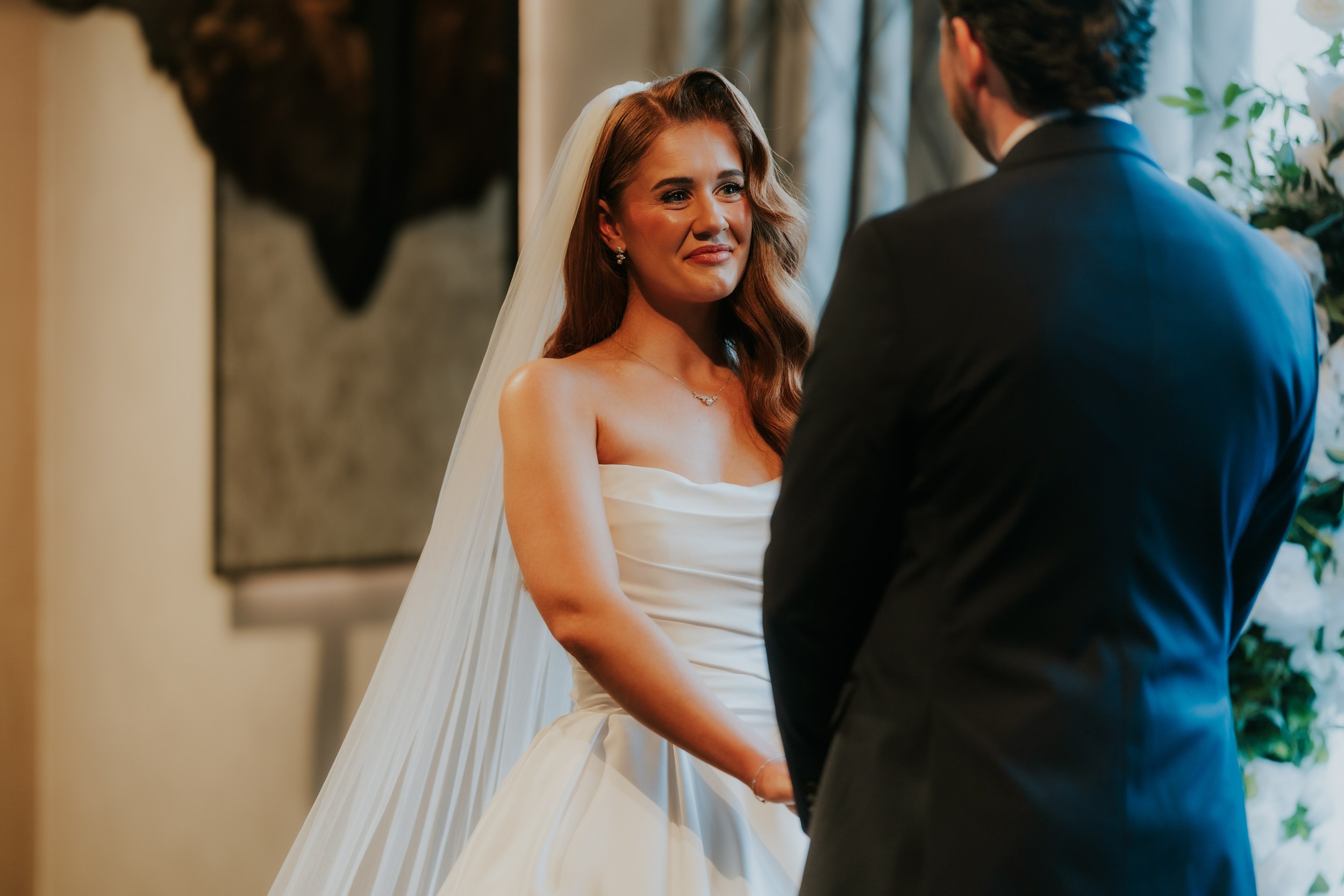 Bride in strapless dress and veil smiling at groom during indoor ceremony