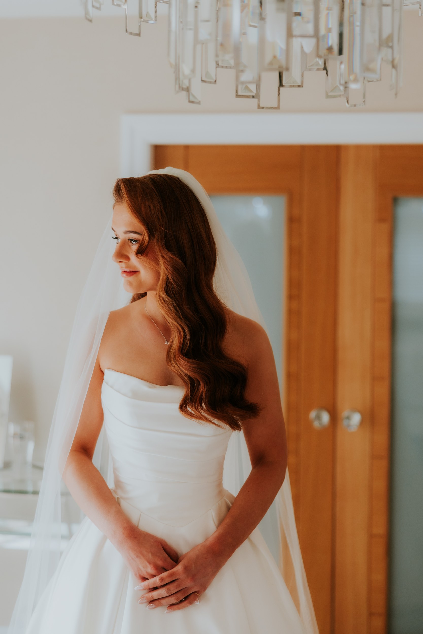 Bride in strapless gown and veil, hands clasped, looking left with wooden doors and chandelier behind.