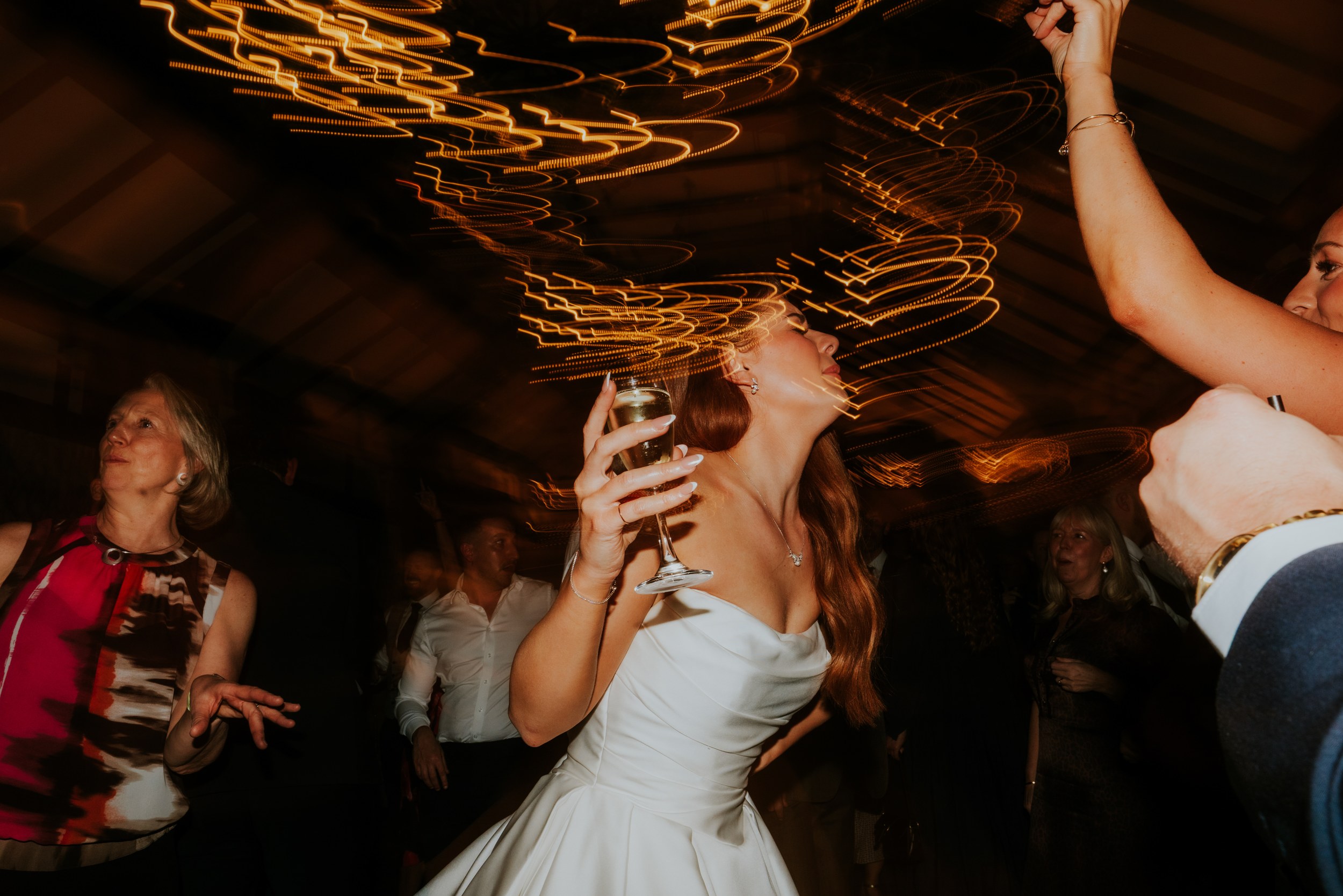 Bride in strapless white dress holding a champagne glass, dancing with guests beneath streaked warm ceiling lights.