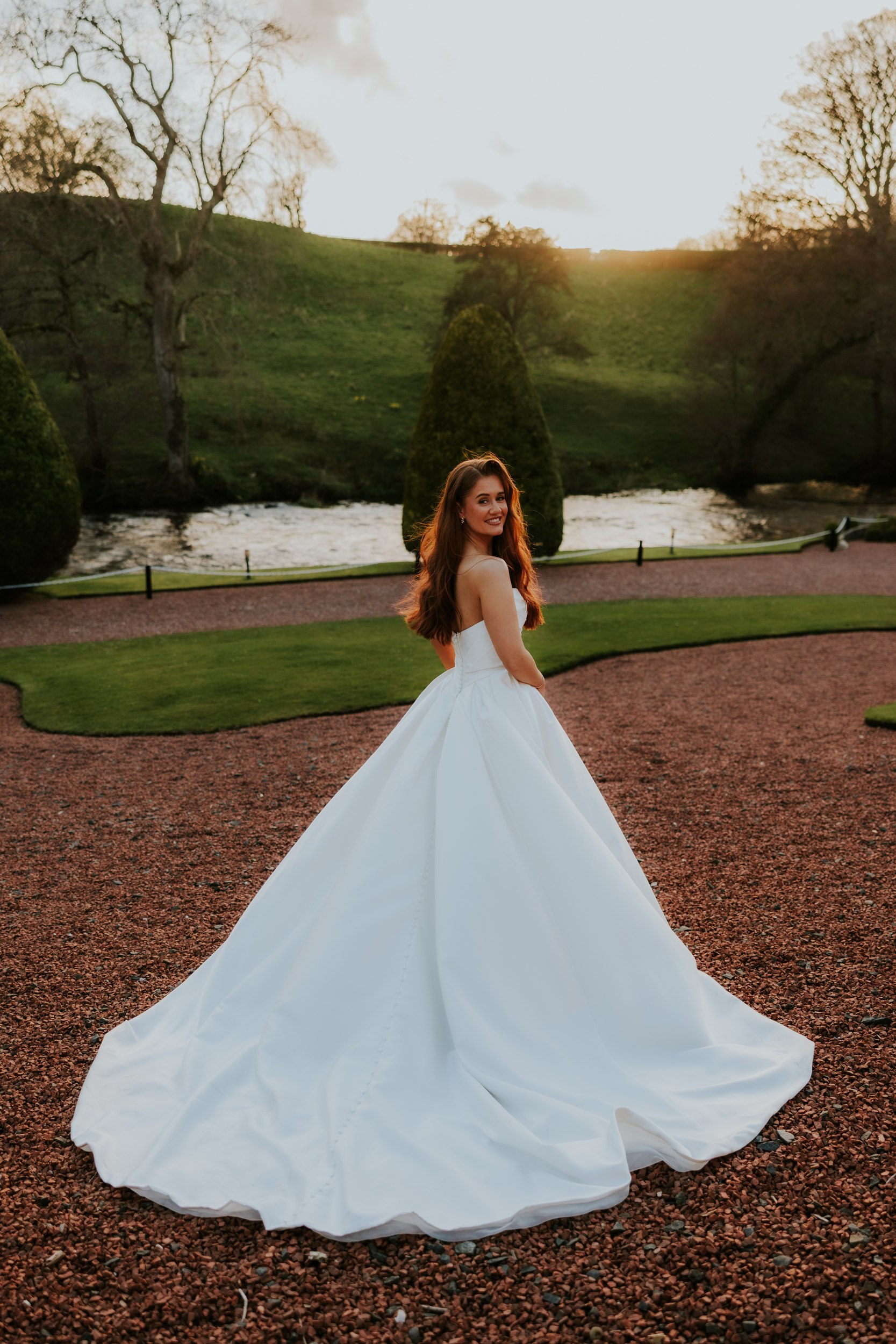 Bride in strapless white gown looking back on a gravel path by a river in hotel gardens at sunset.