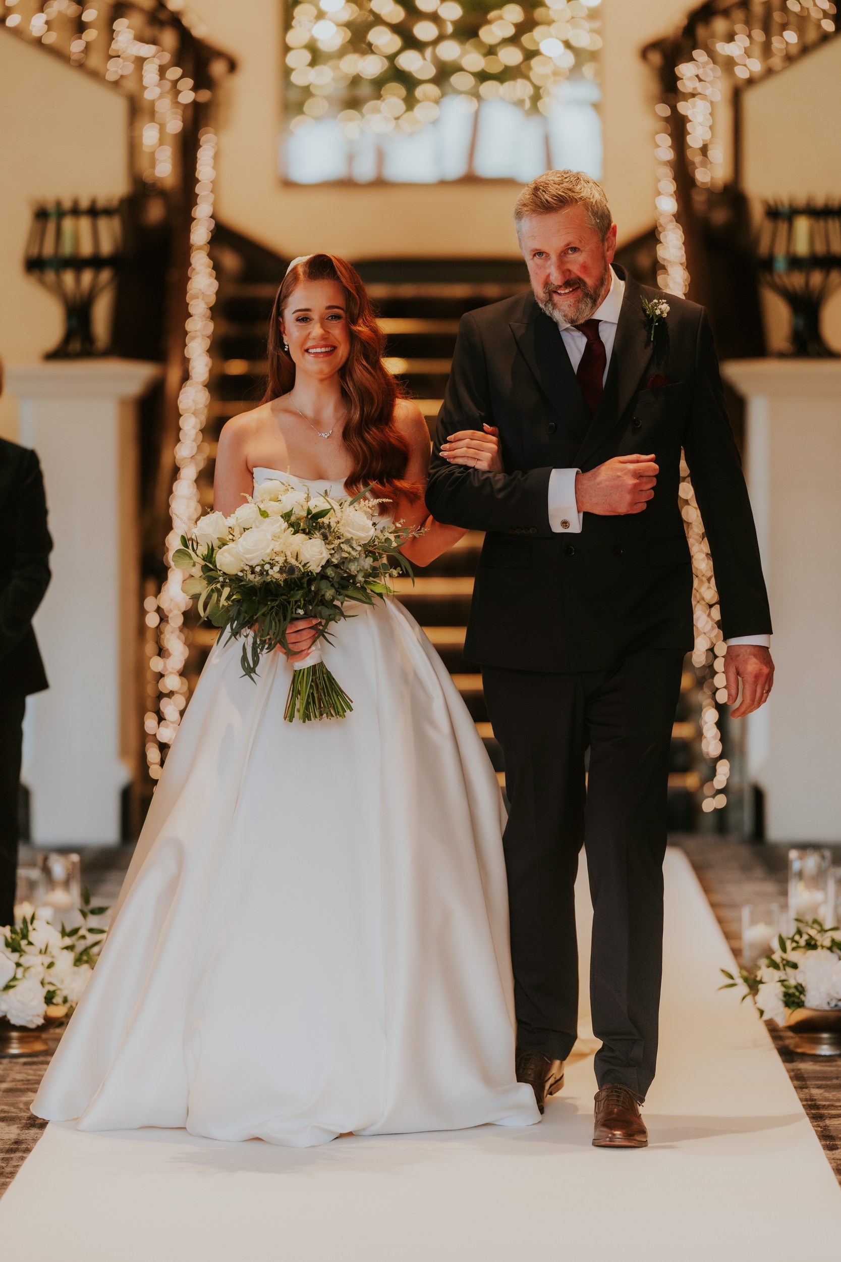 Bride in strapless white gown with bouquet walks arm-in-arm with a man in a dark suit down a fairy-lit aisle.