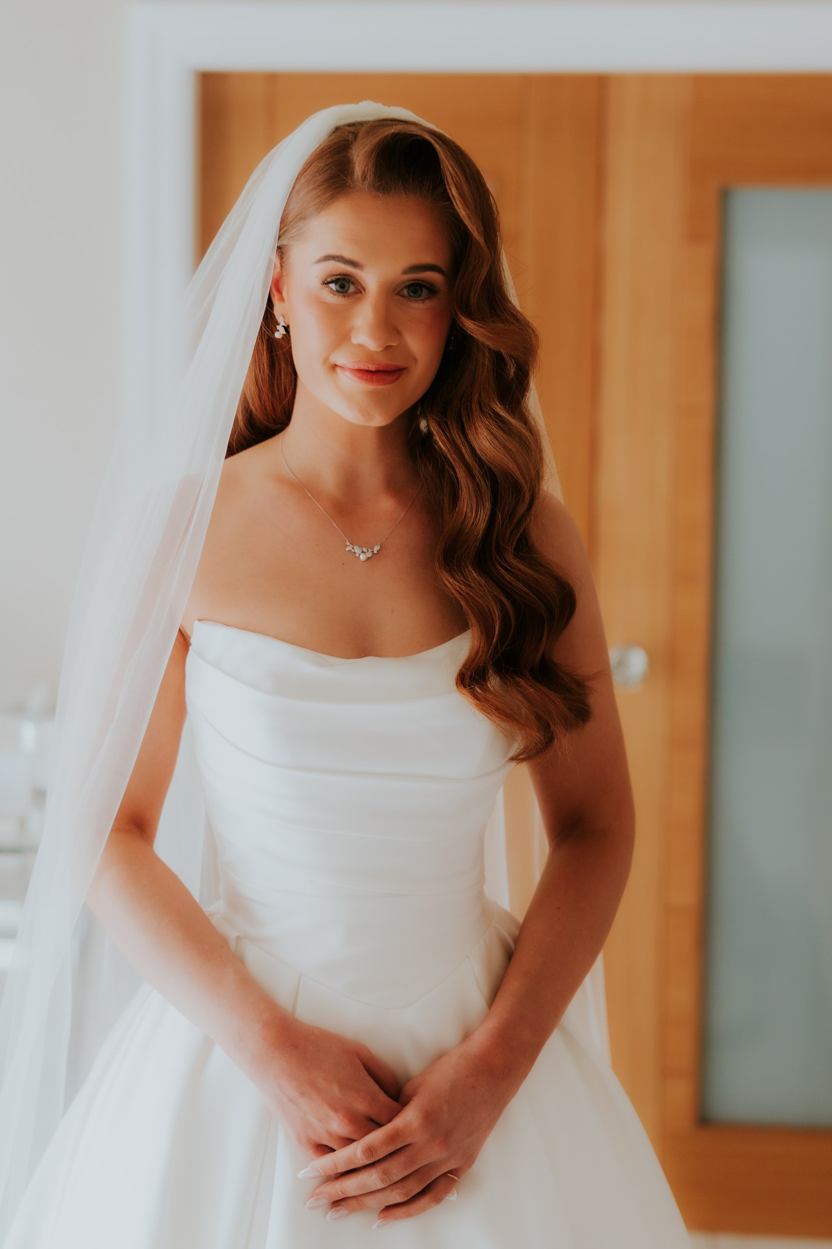 Bride in strapless white wedding dress and veil, side-swept waves, necklace and clasped hands indoors.