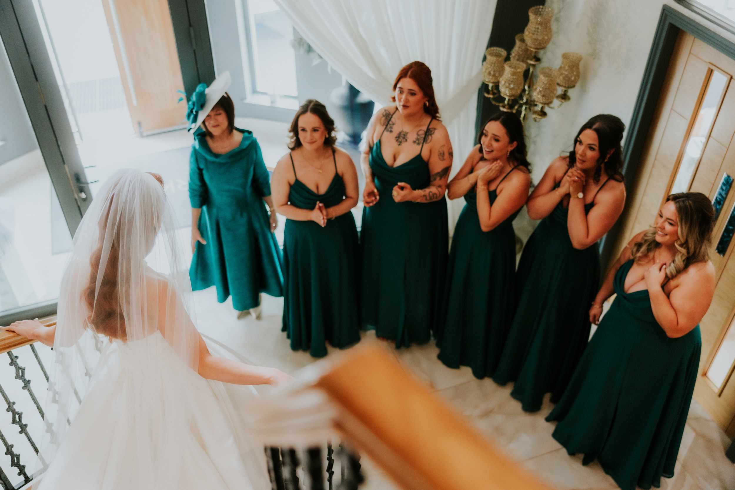 Bride in veil descends staircase as bridesmaids in teal dresses look up, reacting.