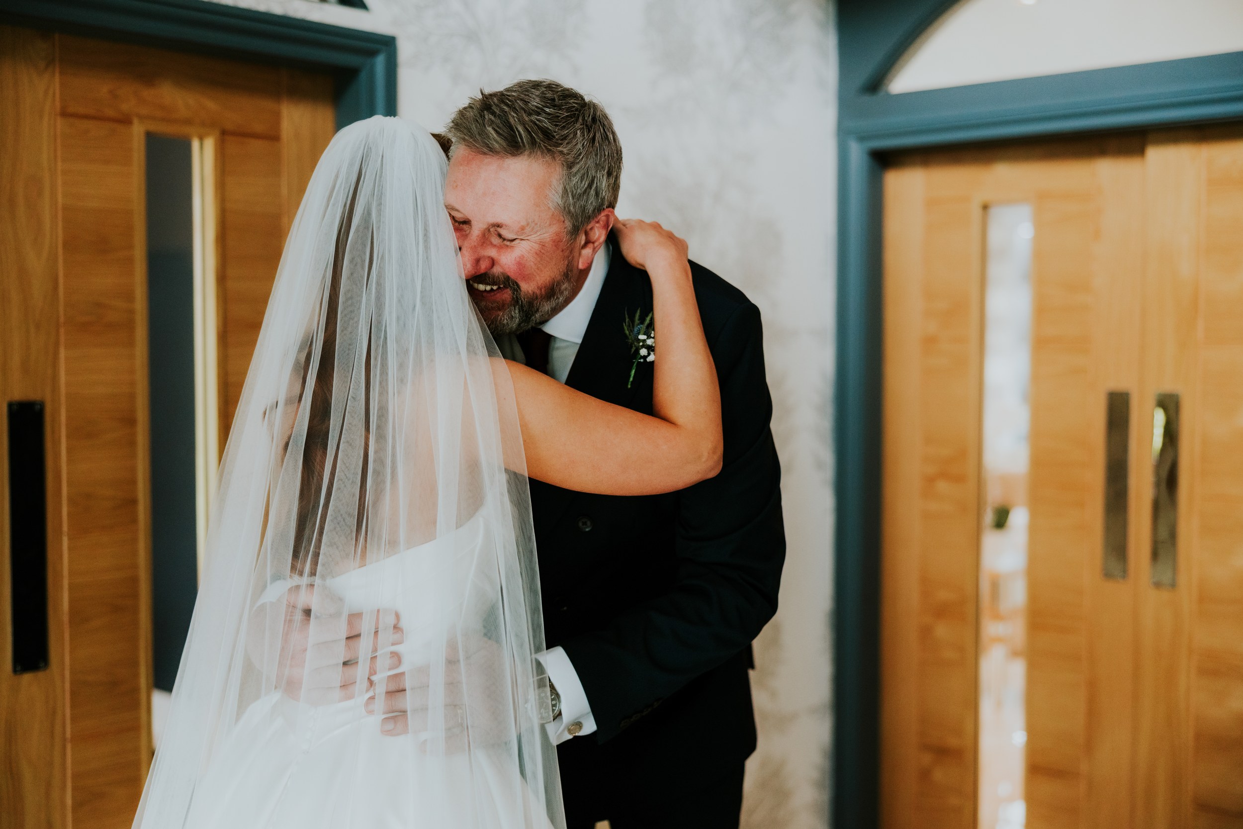 Bride in veil hugging an older man in a suit and boutonniere inside a venue.