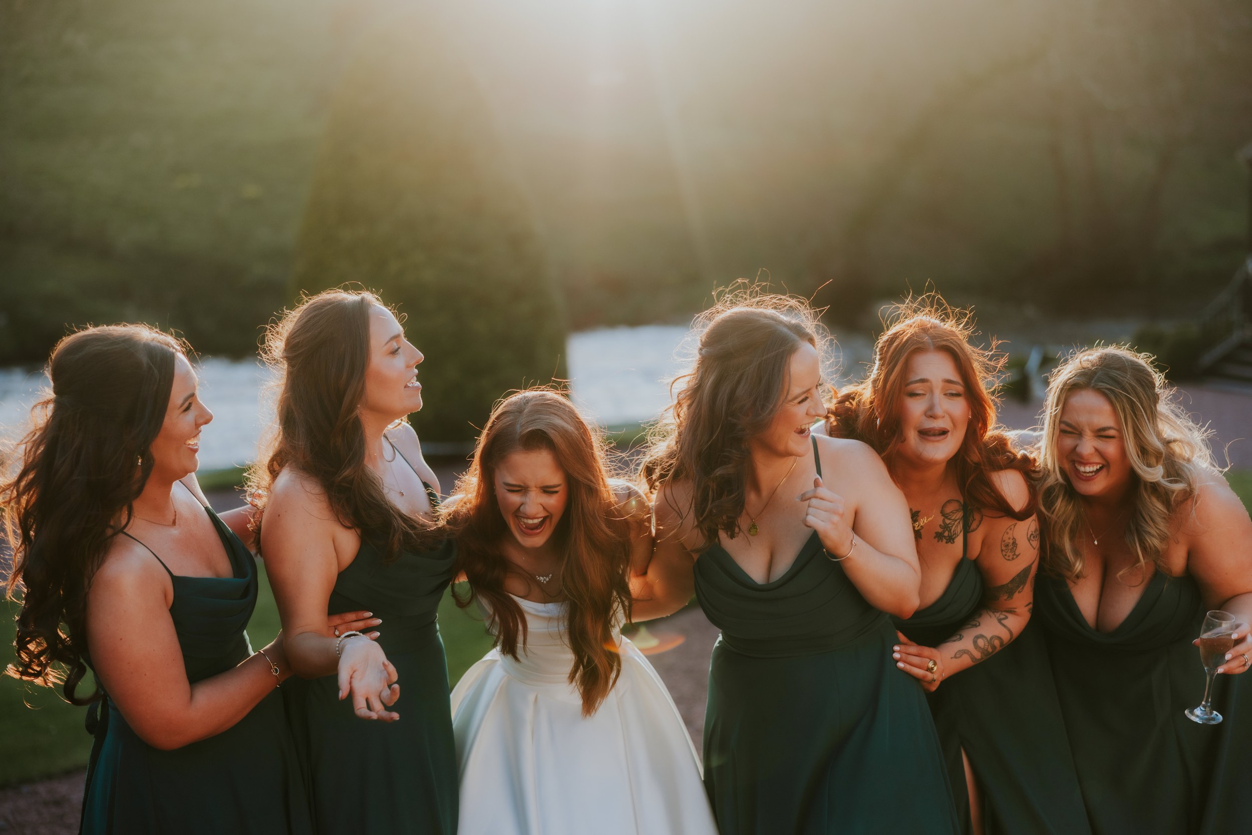 Bride in white and bridesmaids in dark green laughing together outdoors, backlit by low sun.