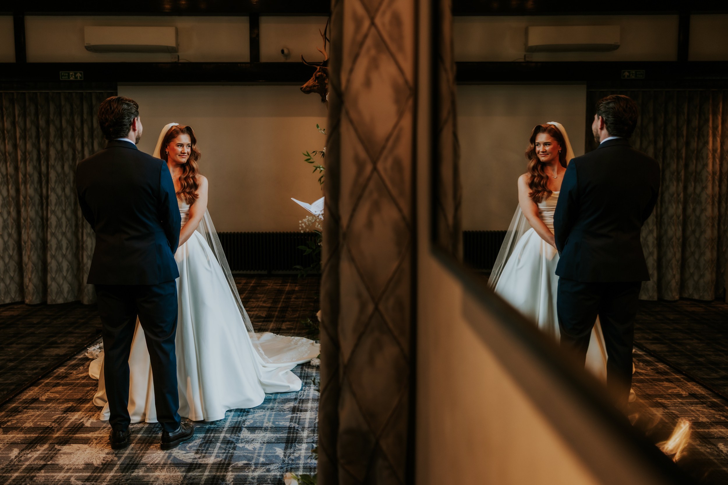 Bride in white dress and veil faces groom inside the venue, their reflection visible in a large mirror.