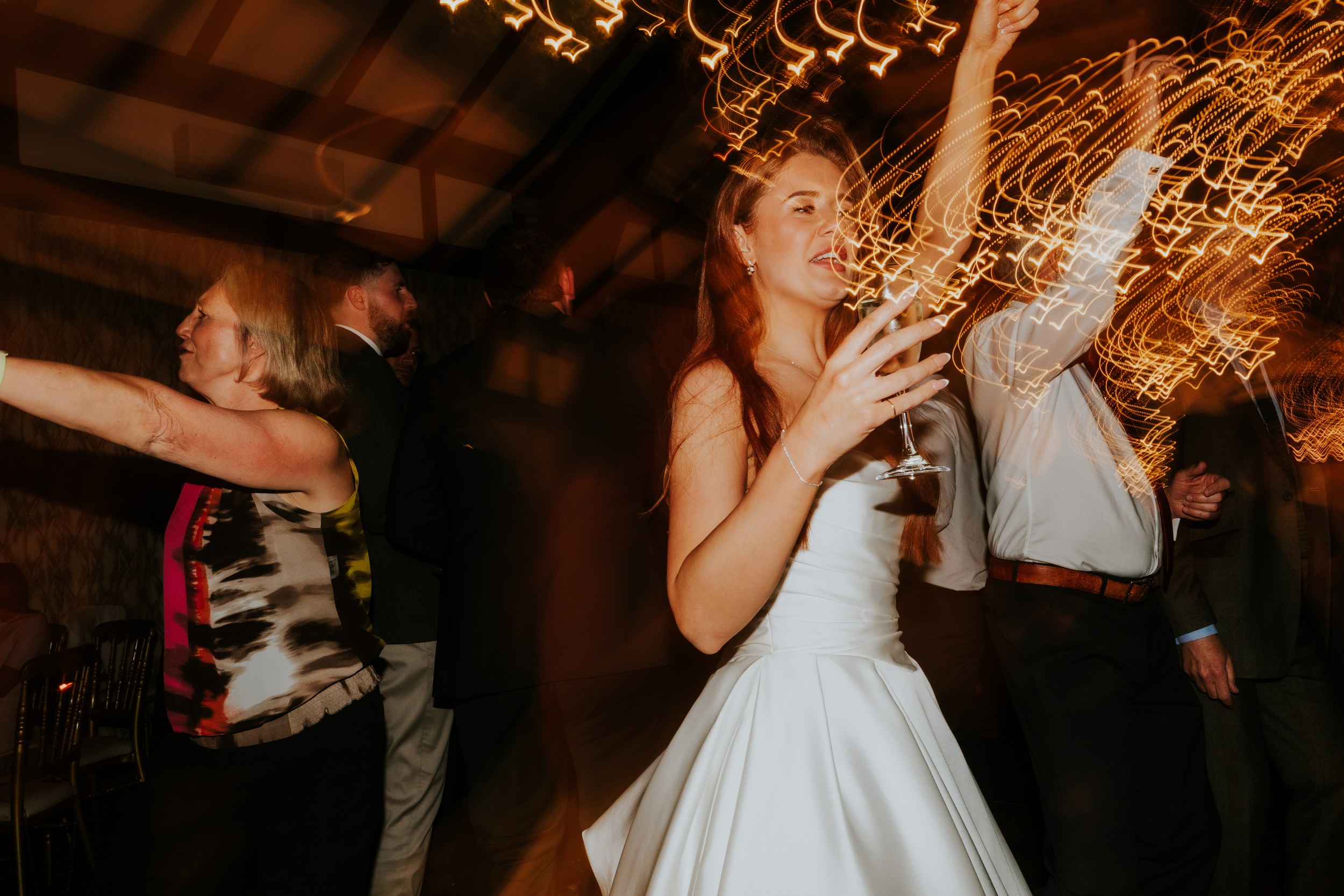 Bride in white dress dancing with a glass amid guests and streaking warm lights.