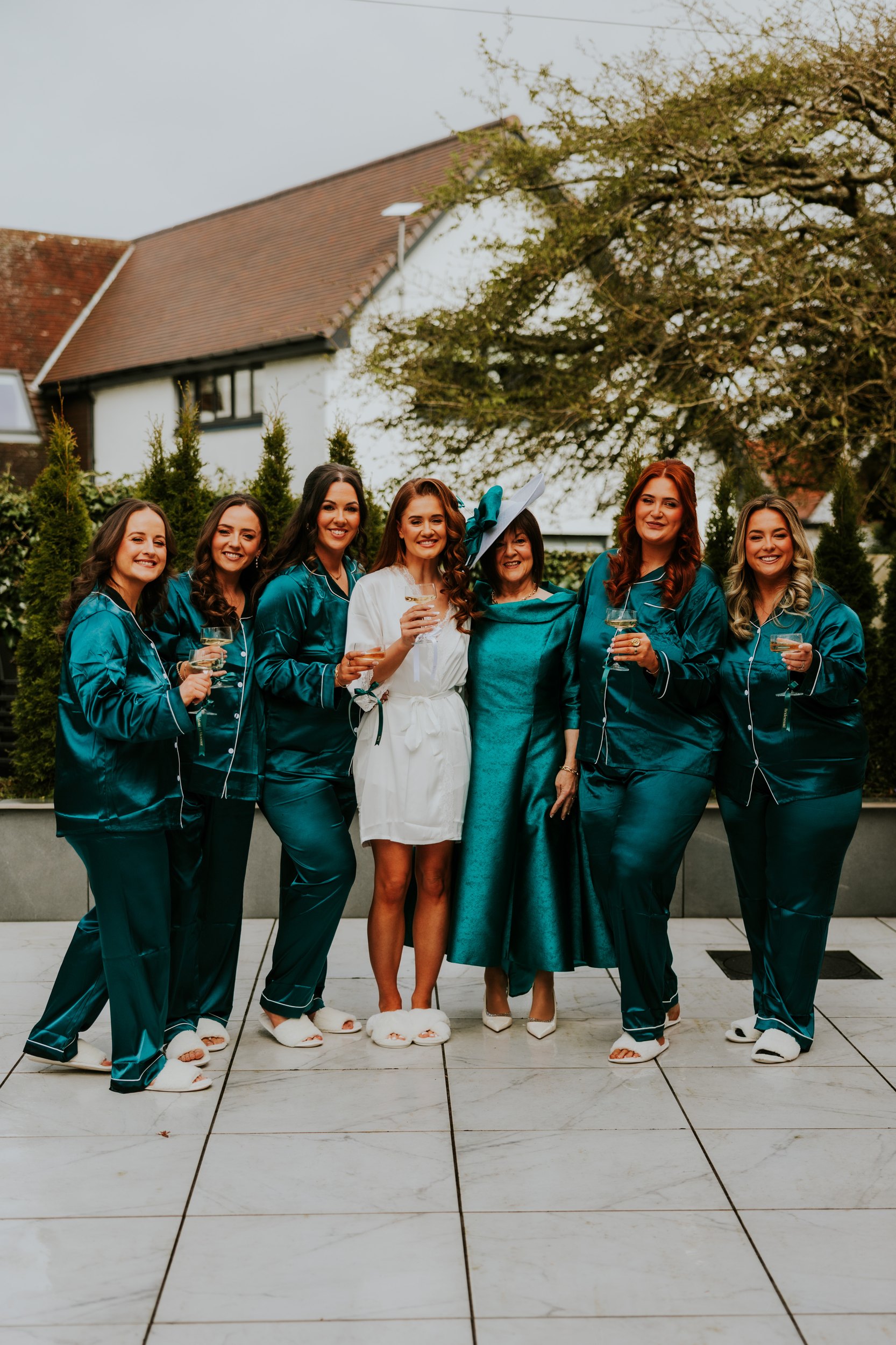 Bride in white robe with bridesmaids in teal satin pyjamas and a woman in a teal dress and hat, holding glasses on a hotel terrace.