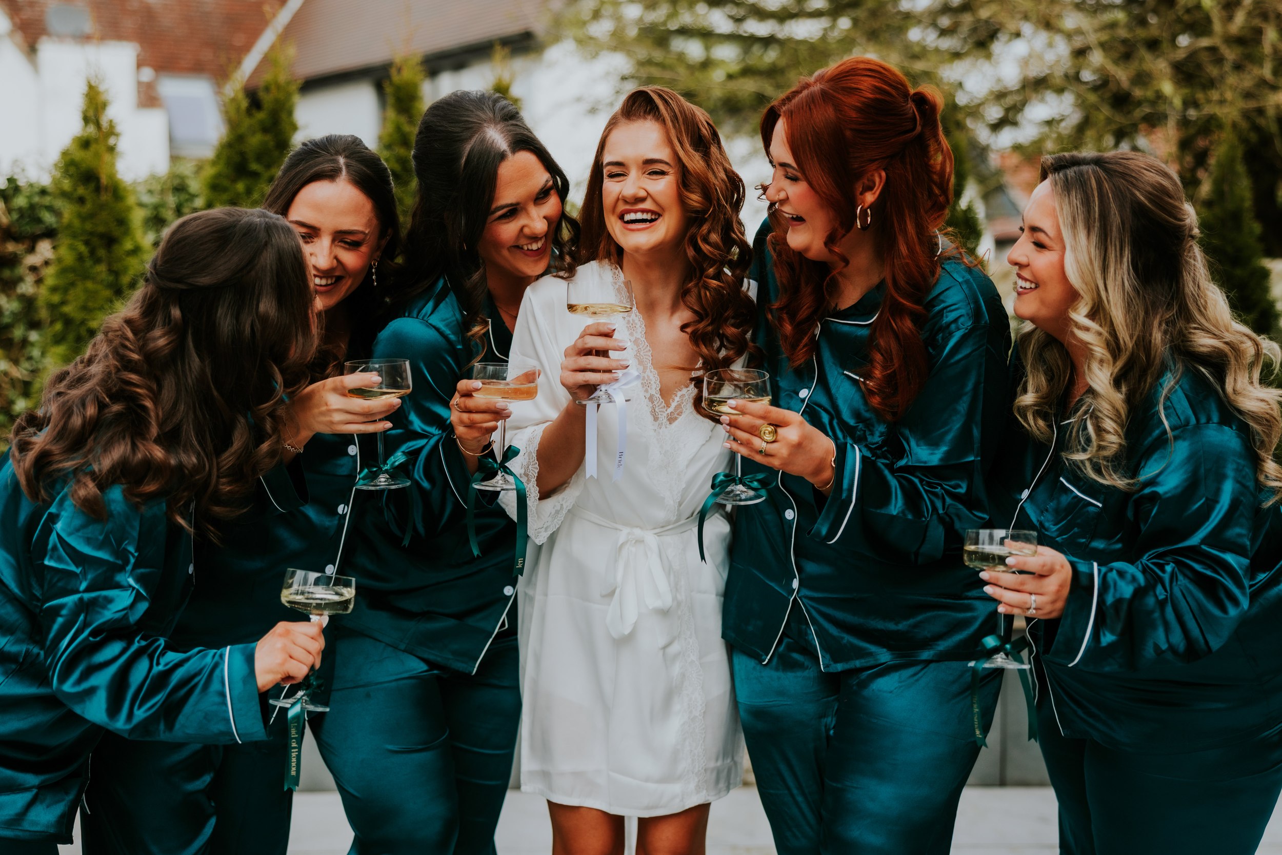 Bride Robyn in a white robe laughing with bridesmaids in teal satin robes holding champagne coupes outdoors.