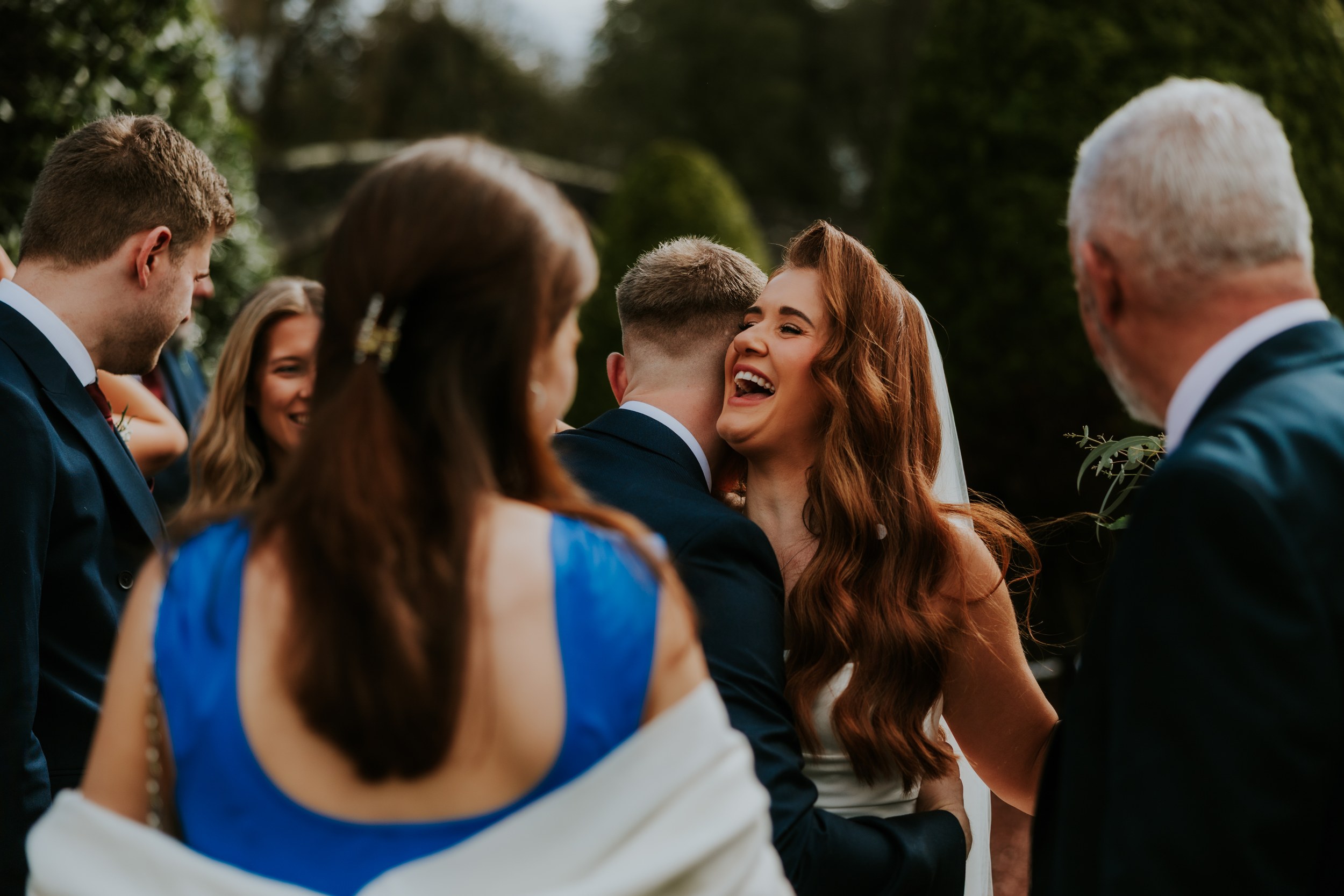 Bride Robyn laughing as she hugs a man in a suit, surrounded by wedding guests outdoors.