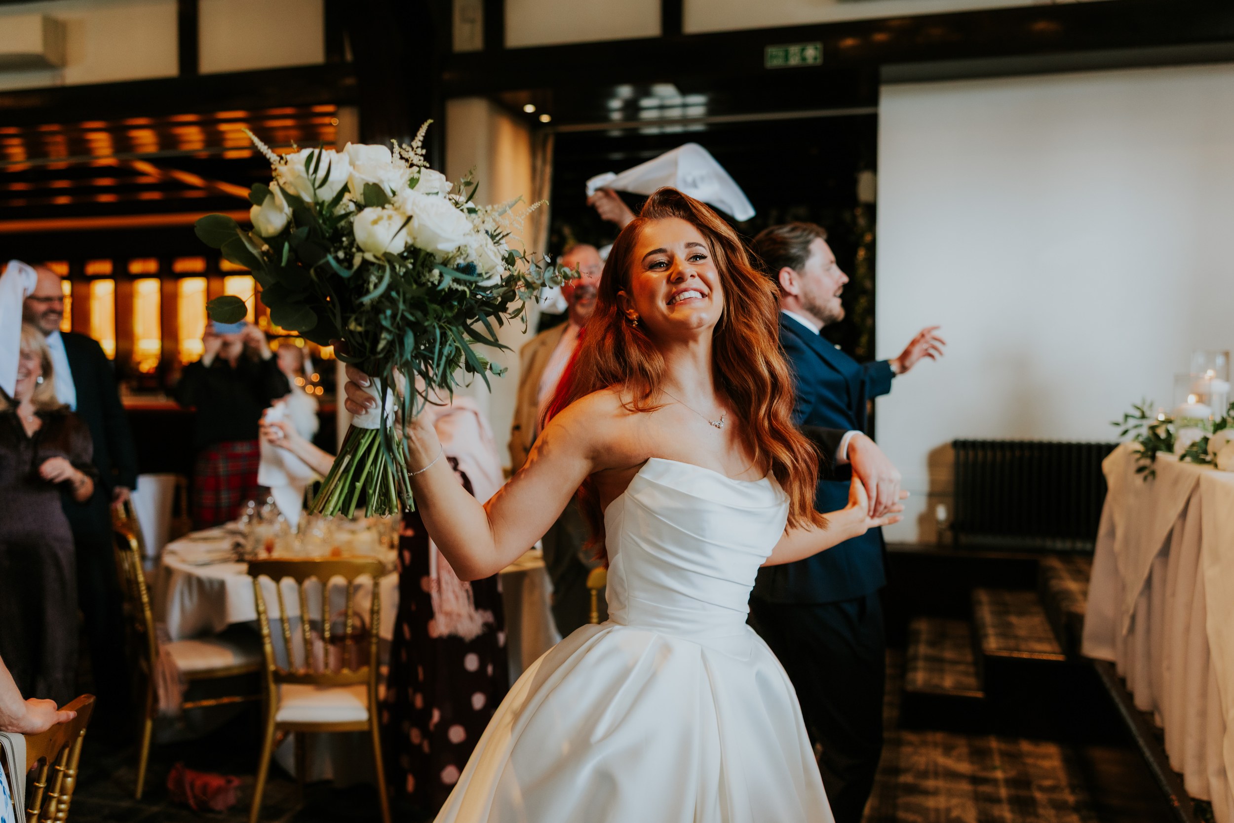 Bride Robyn smiling, holding a large white bouquet in the reception as guests wave napkins.