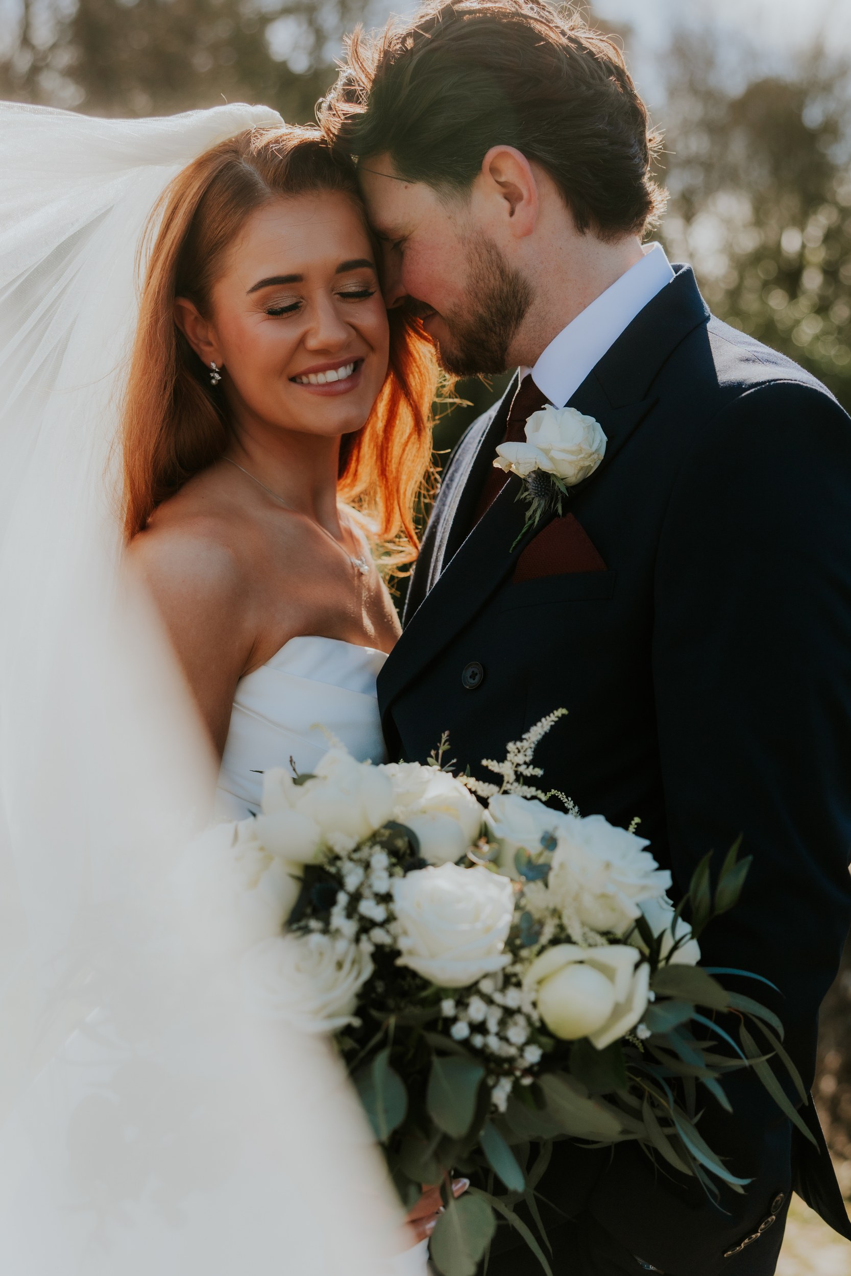Bride Robyn smiling with veil and white-rose bouquet as groom Jack leans in, foreheads touching.