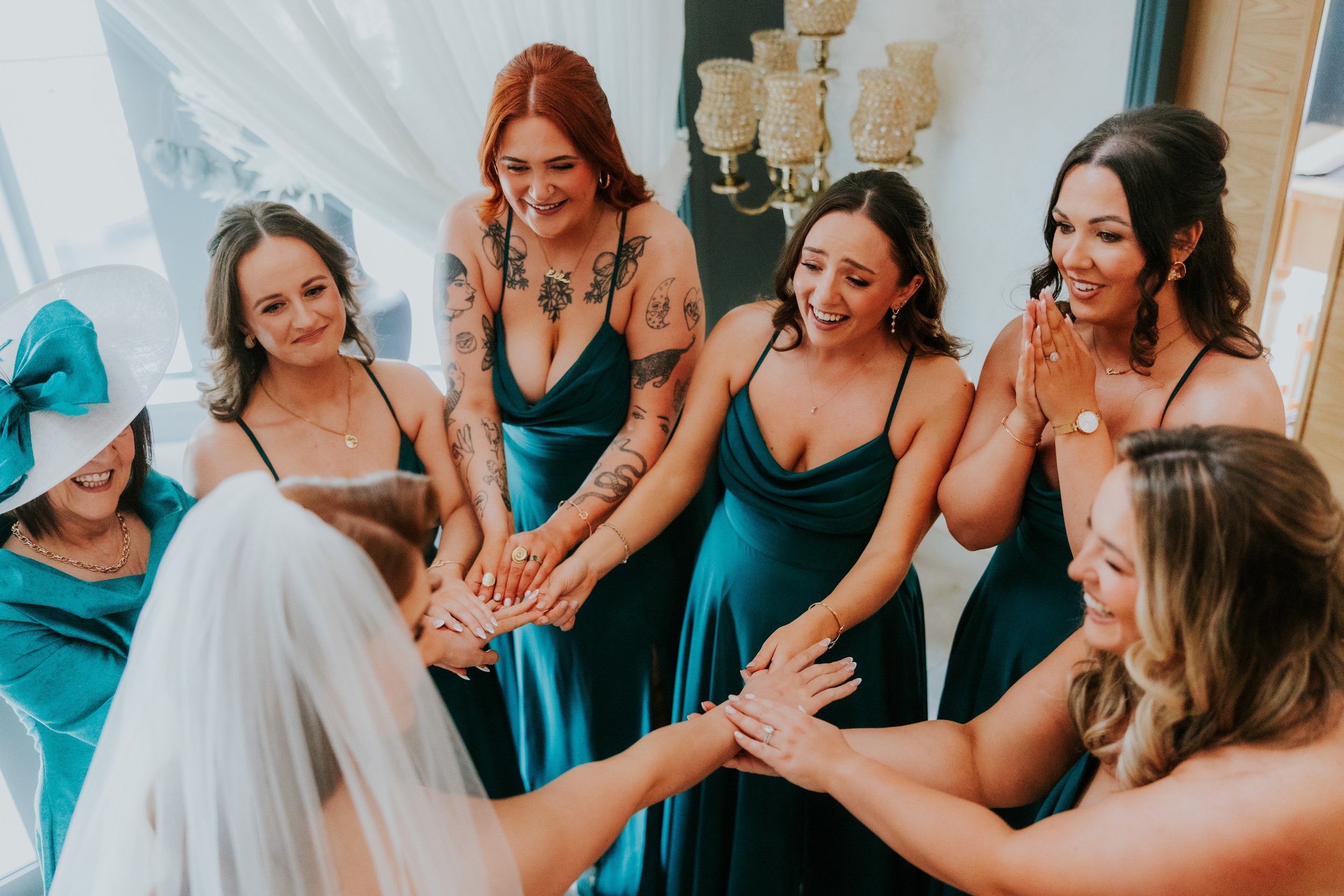 Bride with veil and bridesmaids in teal dresses join hands in a circle, smiling during a pre-wedding moment.