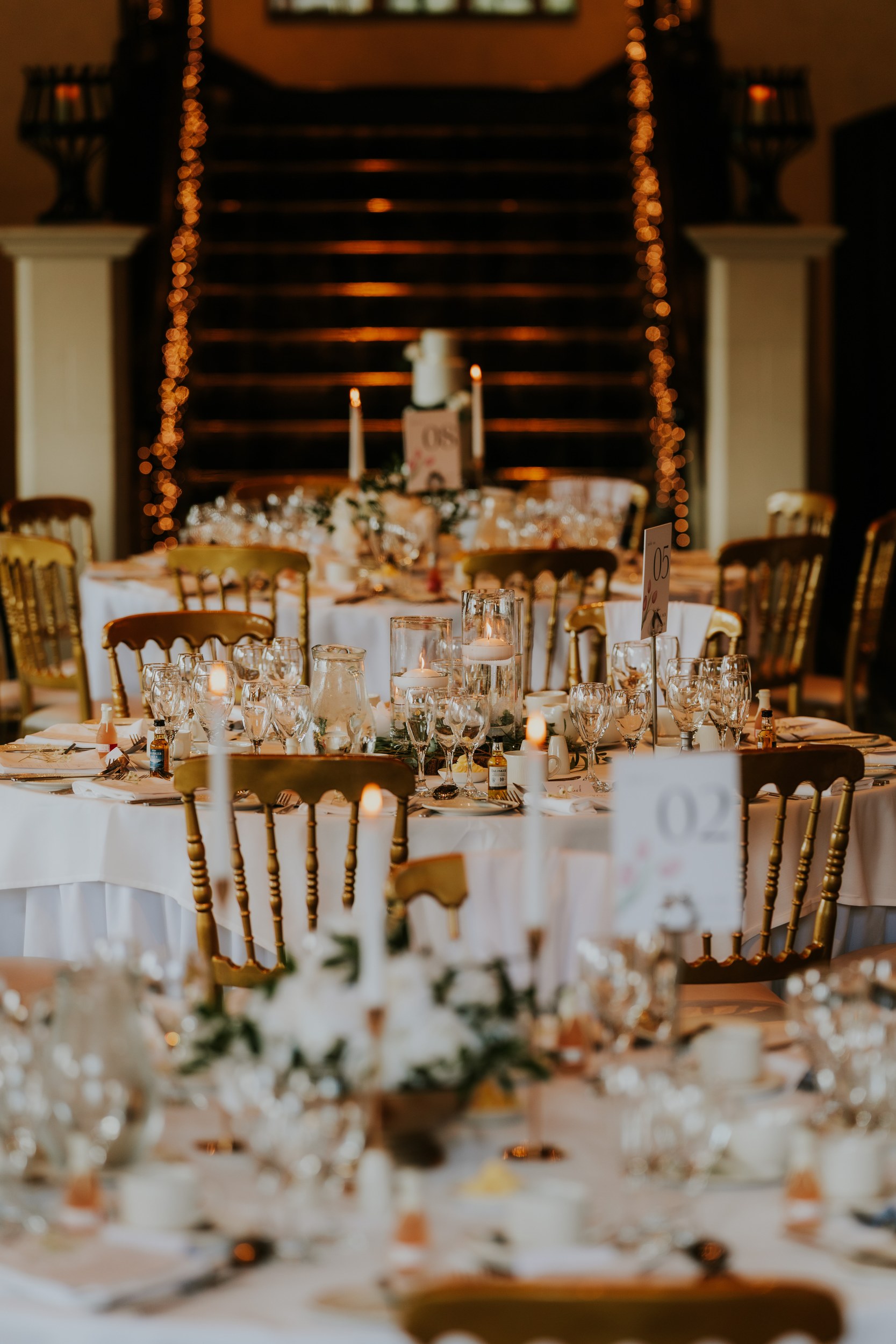 Candlelit wedding reception tables with glassware, floral centrepieces and gold chairs, fairy lights on a staircase behind.