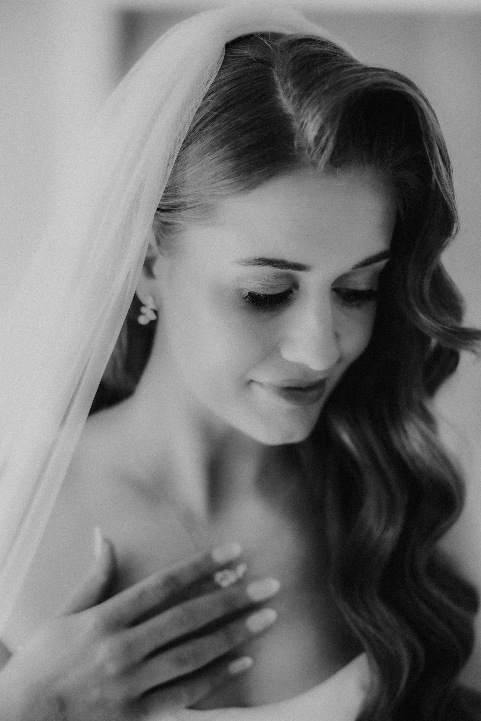 Close-up black-and-white portrait of bride Robyn in a veil, looking down with her hand on her chest.