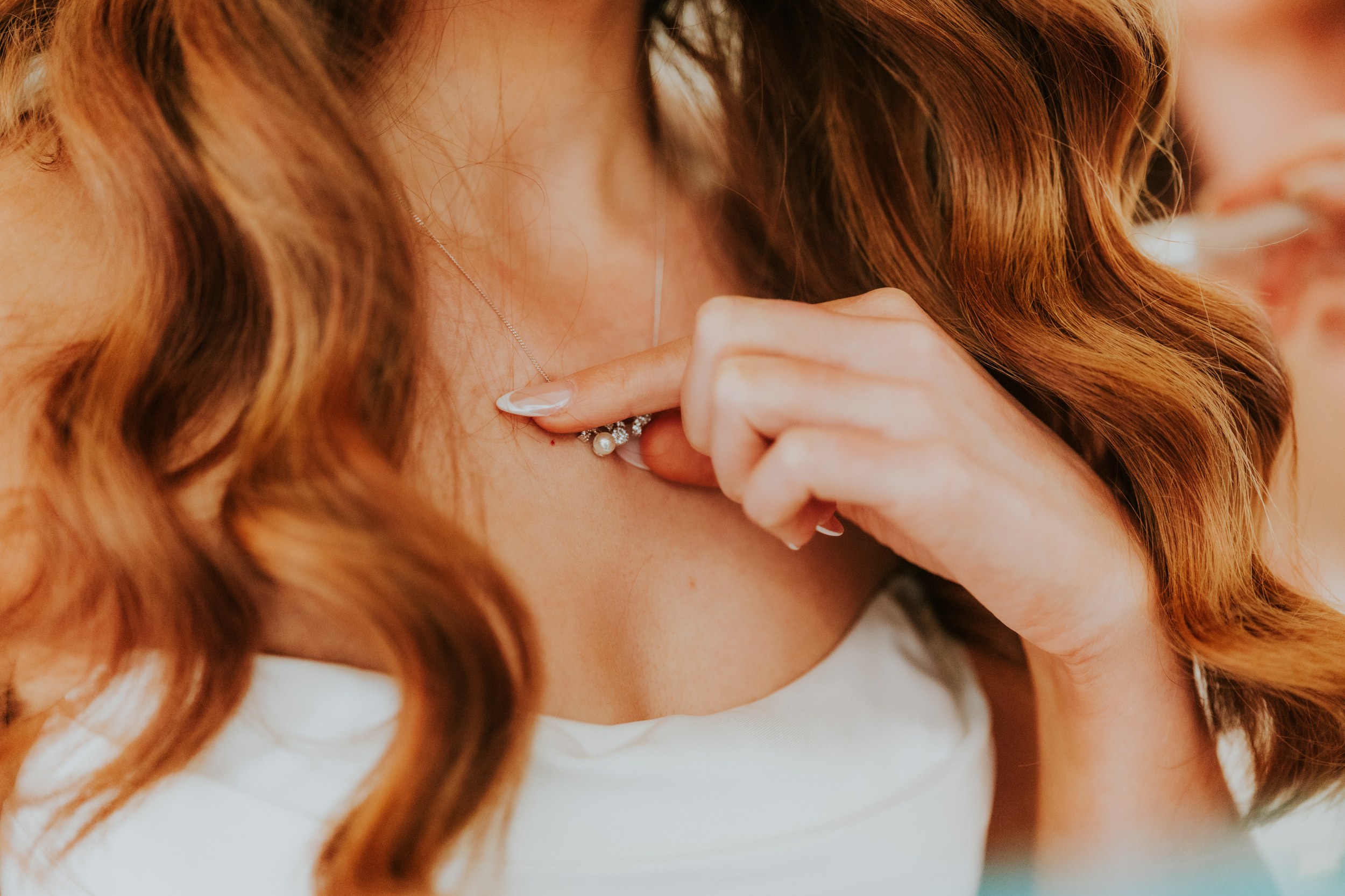 Close-up of bride fastening a pearl-and-diamond pendant, wavy auburn hair and a French-tip manicure visible.
