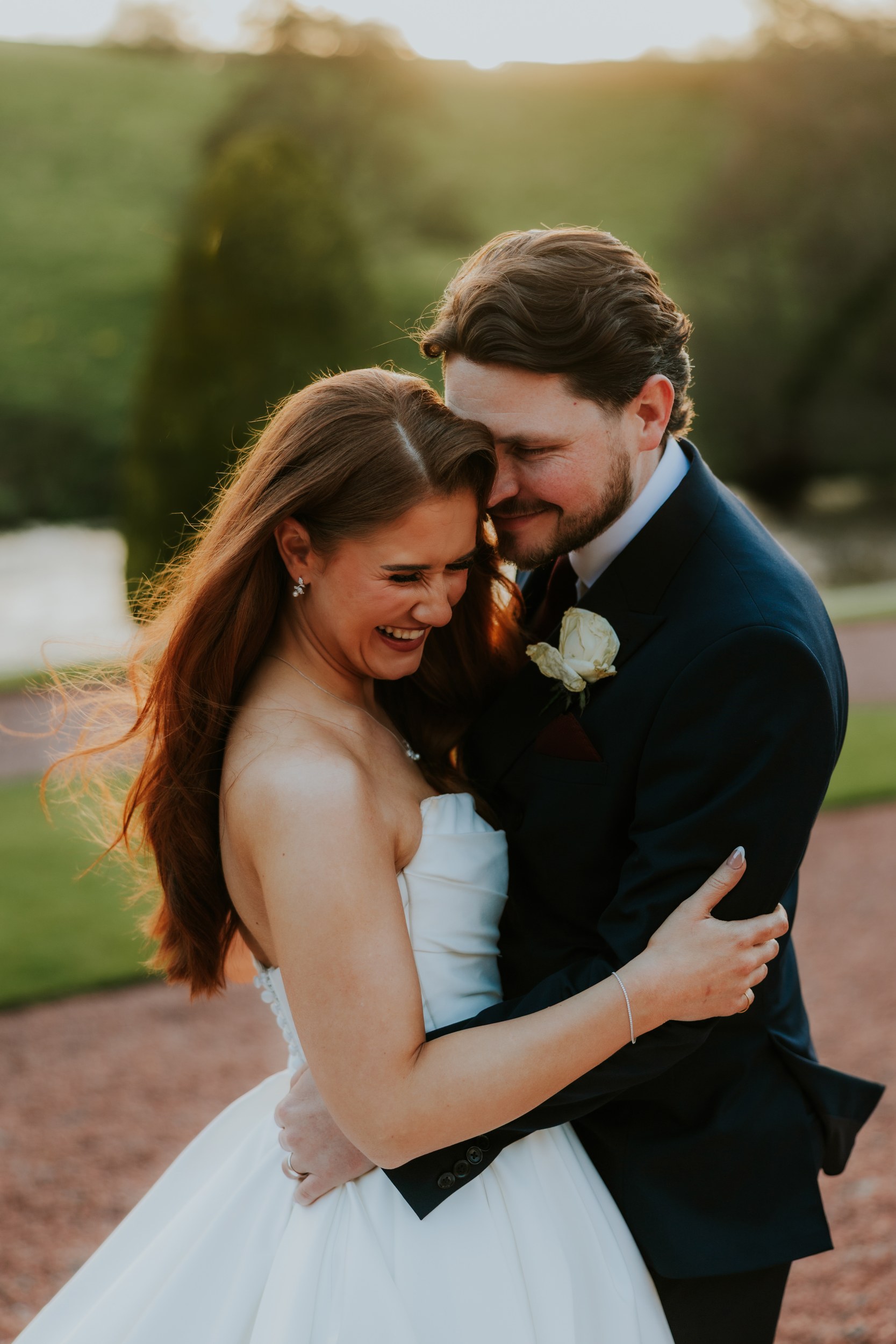 Couple laughing in an embrace outdoors, bride in a strapless white dress and groom in a dark suit with a white rose boutonniere.
