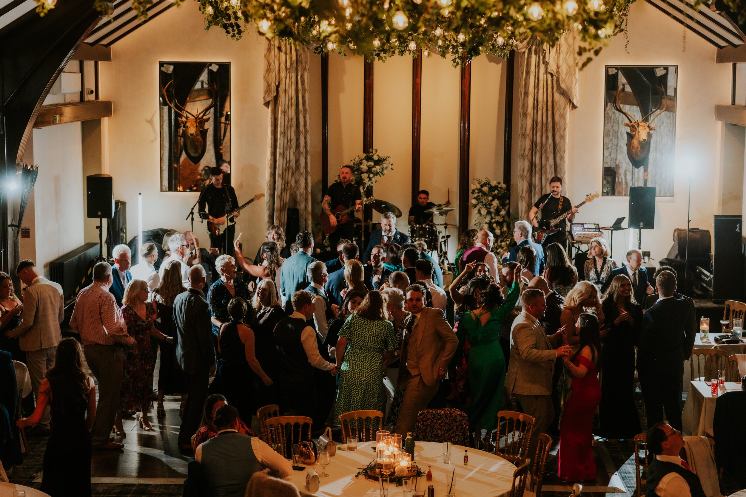 Crowd dancing at a wedding reception with a live band on stage under hanging greenery and candlelit tables.