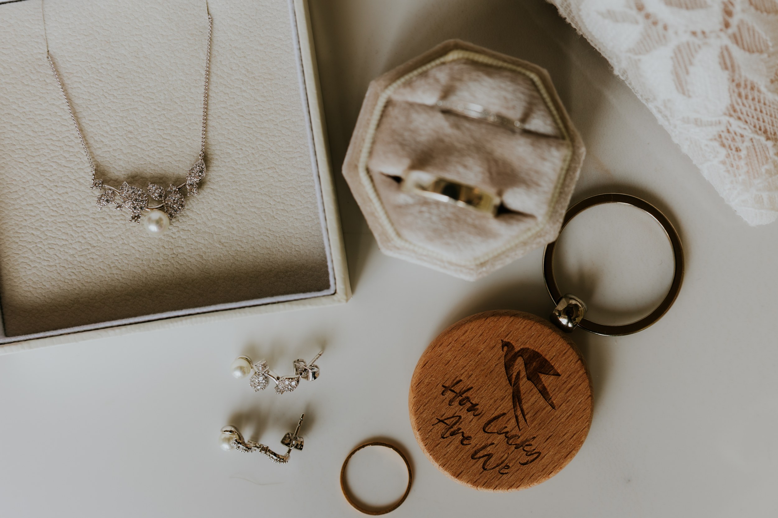 Flat lay of bridal jewellery: pearl-and-crystal necklace, pearl drop earrings, gold wedding rings and velvet ring box beside lace.
