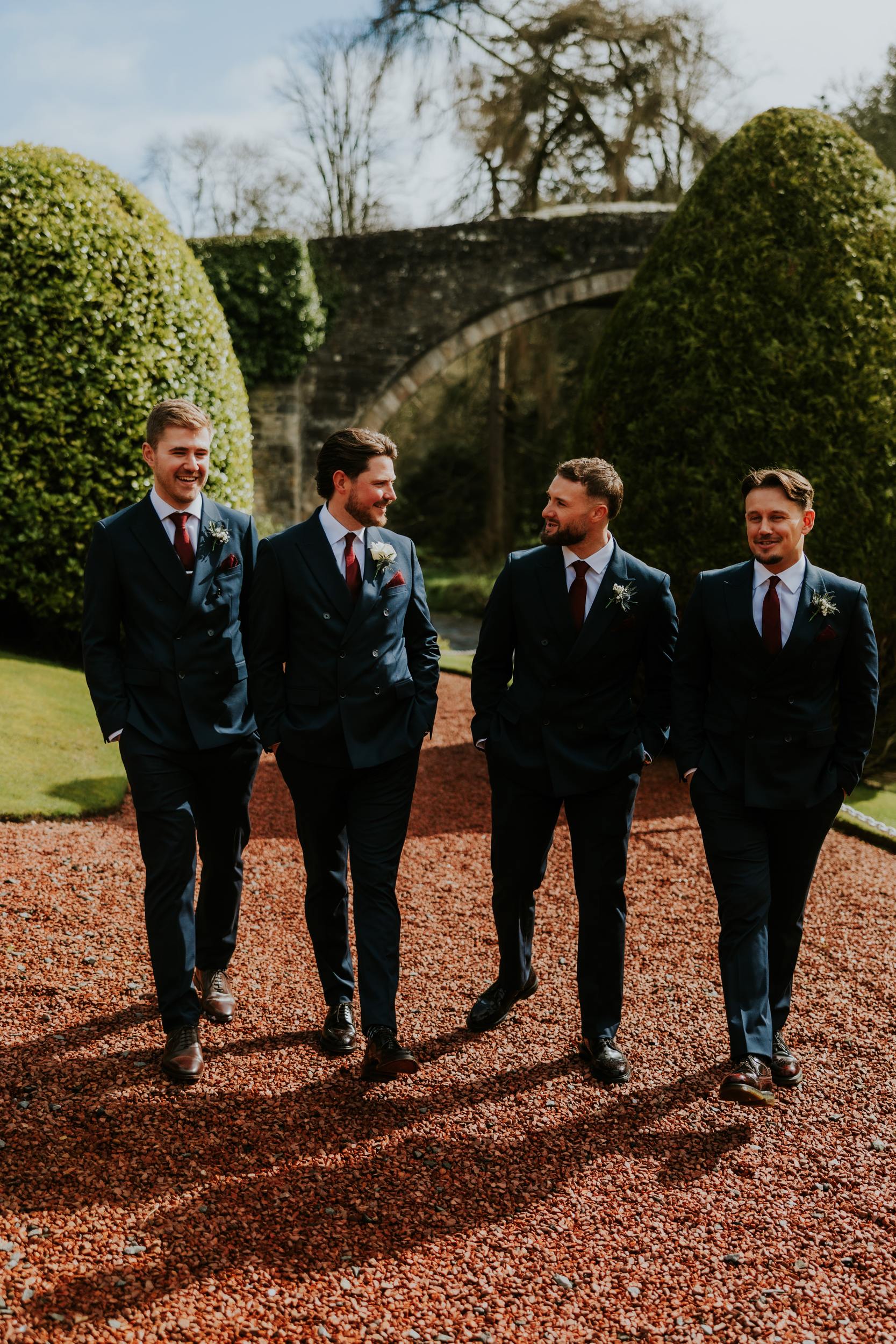 Four groomsmen in navy suits walking on red gravel path with clipped yew hedges and a stone bridge arch.