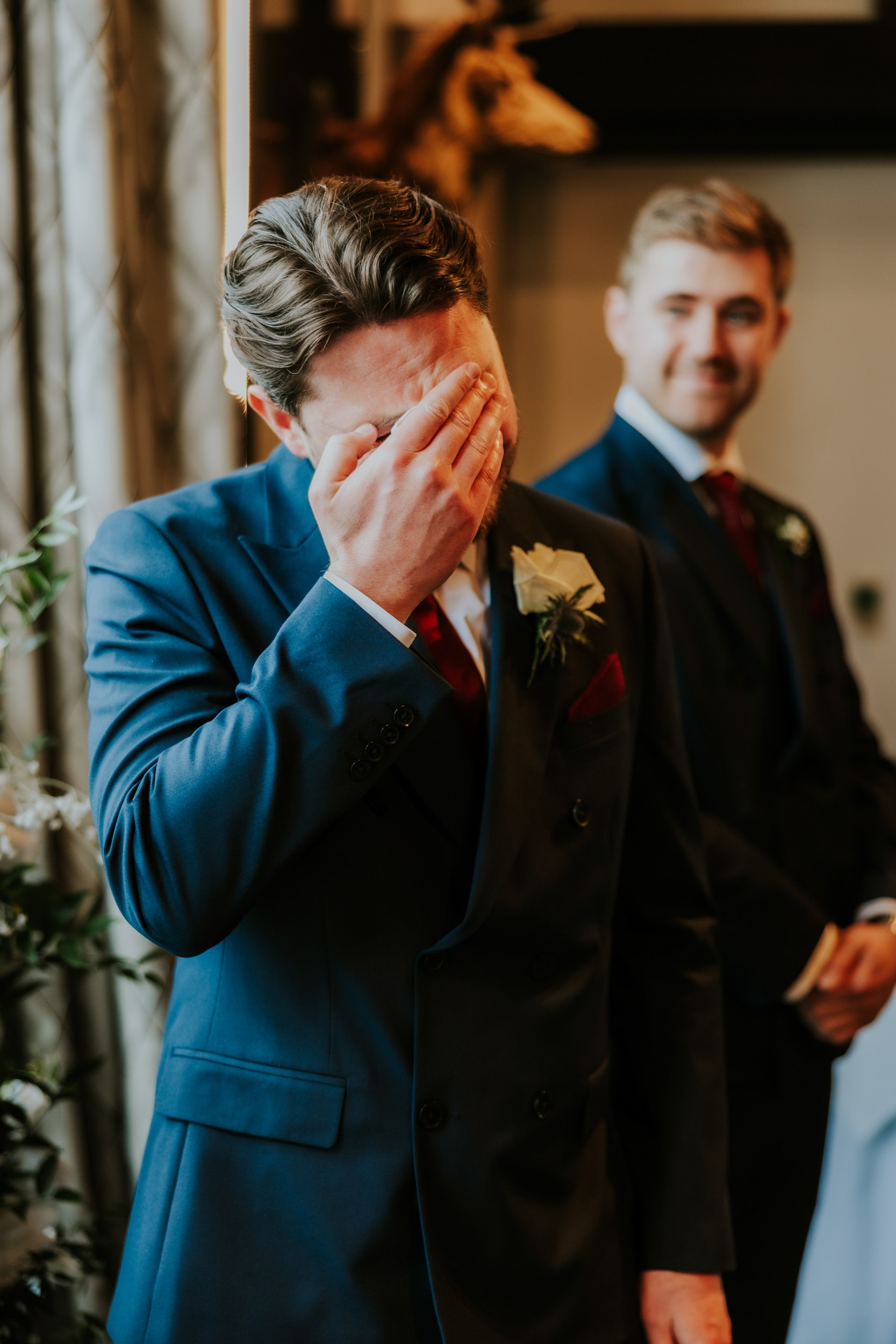 Groom in a navy suit covers his face with his hand while a smiling groomsman stands behind him at an indoor wedding.